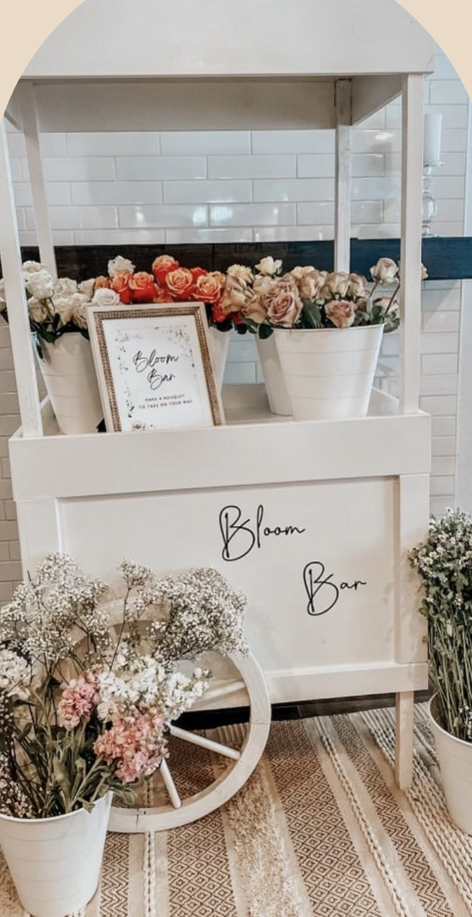 A display of flower arrangements in white pots on a blooming bar stand, with a framed sign that says 'Bloom Bar' and a variety of pink, peach, and white flowers. Additional flowers are in a white vase on a patterned rug.