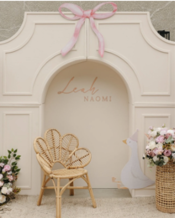 Decorative wall with pink bow, rattan chair, flowers, and a paper cutout of a swan wearing a pink hat and bow.