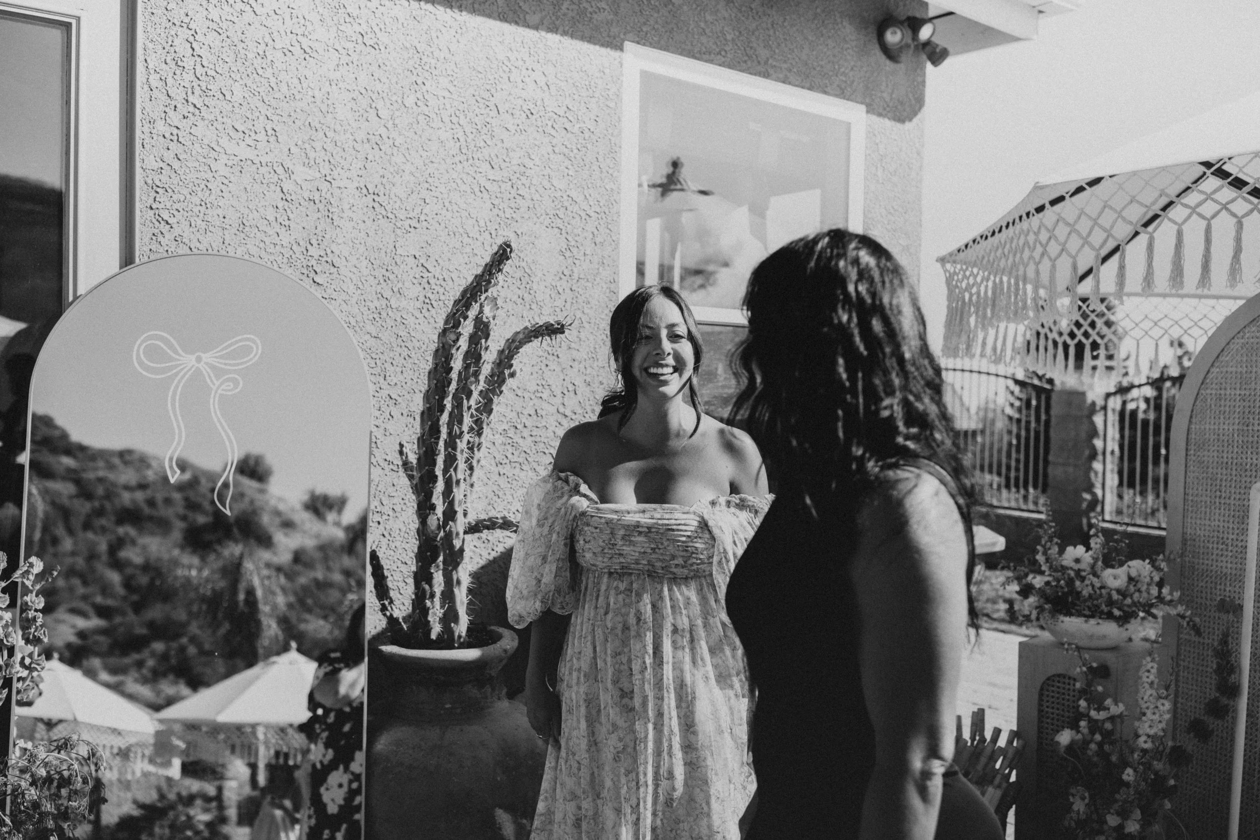 Two women talking on a terrace, one smiling and wearing an off-the-shoulder dress, the other with long dark hair wearing a dark sleeveless top, outdoor patio setting with a cactus in a pot and parasol.
