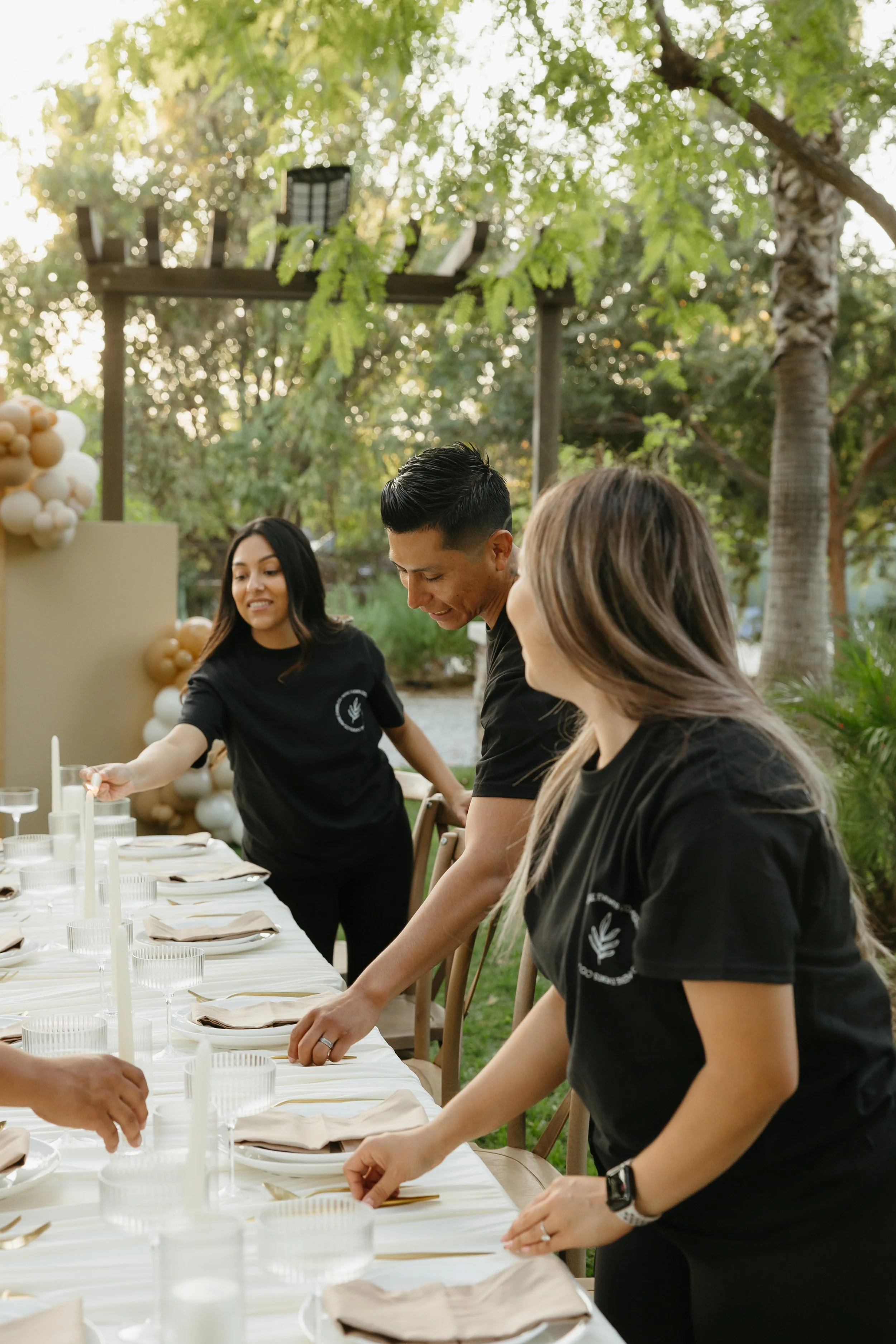 People setting a table outdoors with greenery and trees in the background, during daytime.