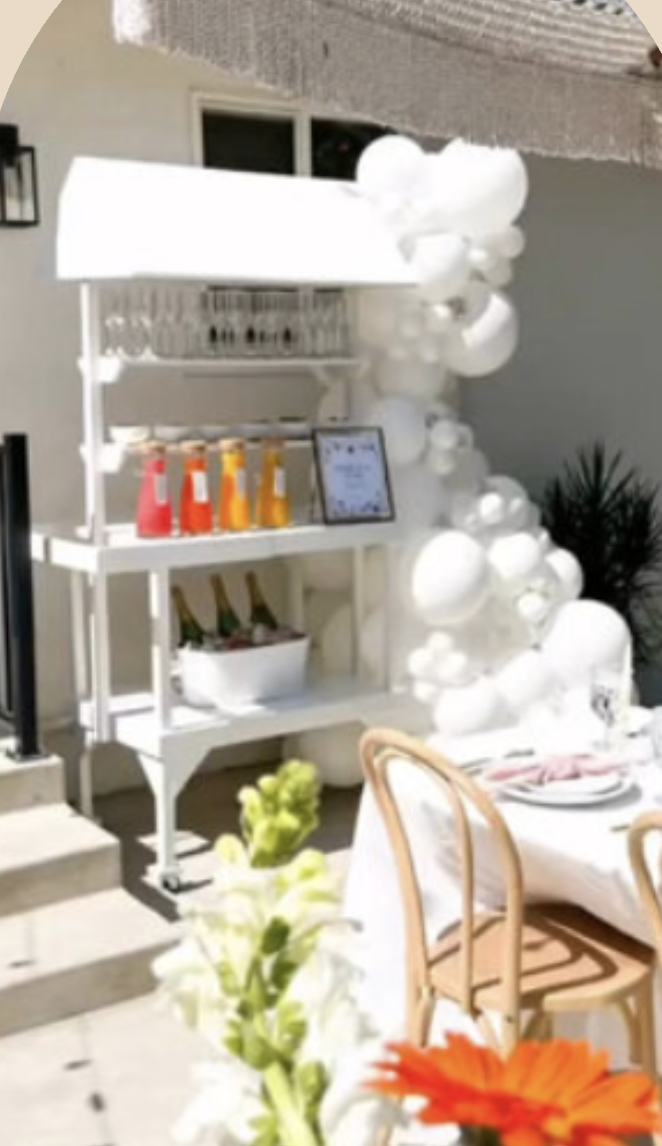 Outdoor party setup with balloon decorations, a white cart with colorful drinks, a table with place settings, and orange and white flowers.
