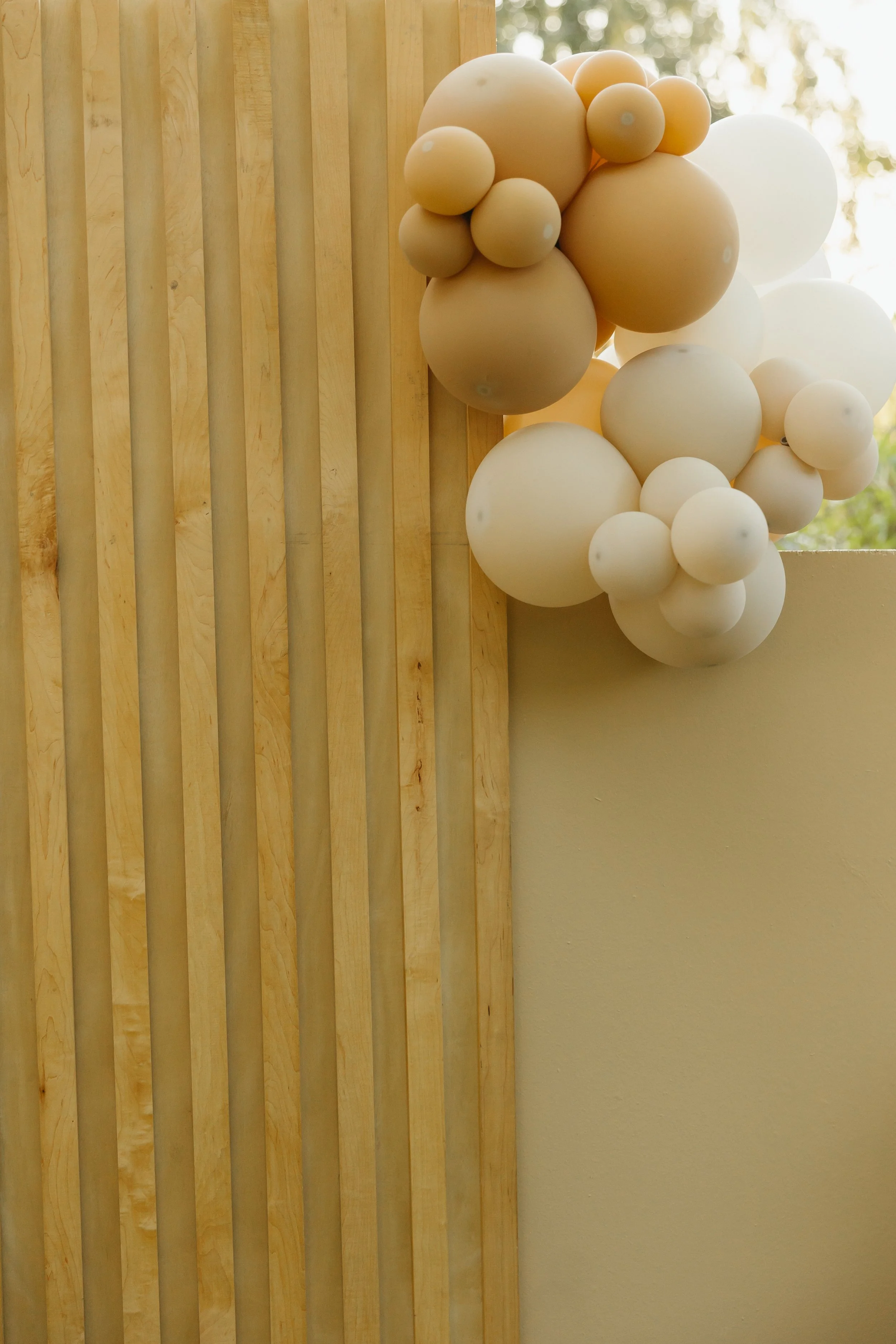 Cream-colored balloons arranged in clusters, attached to a wooden vertical slat wall, with a partial view of a white wall and blurred outdoor scenery in the background.