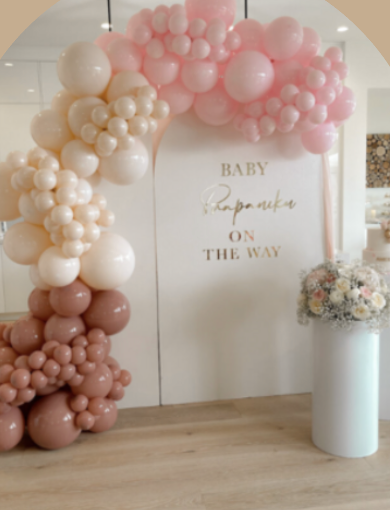 Decorative balloon arch with pink, white, and peach balloons and a sign that says "Baby Propamake on the way," with a bouquet of flowers in a white vase nearby.