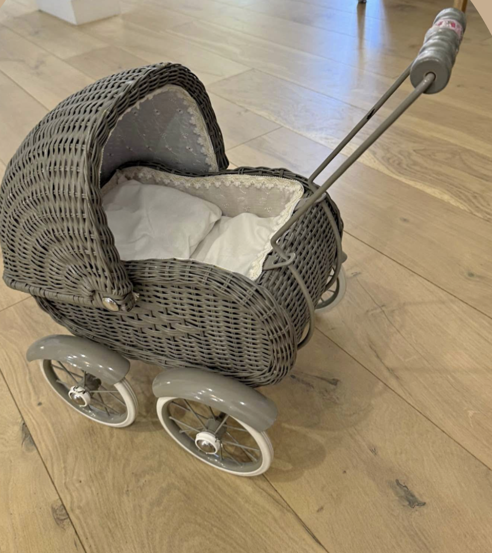 A small vintage-style toy stroller with a wicker design, white wheels, and a cushion lined interior sitting on a wooden floor.