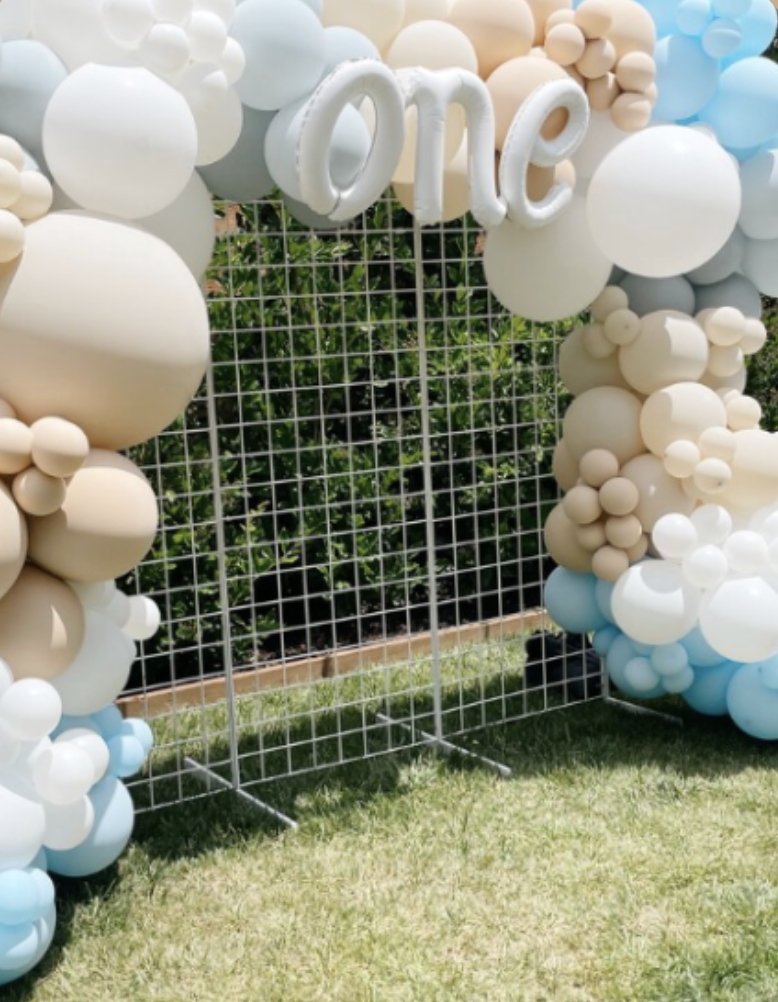 Decorative balloon arch with the word 'one' in white letters, set outdoors on a grassy area with greenery in the background.