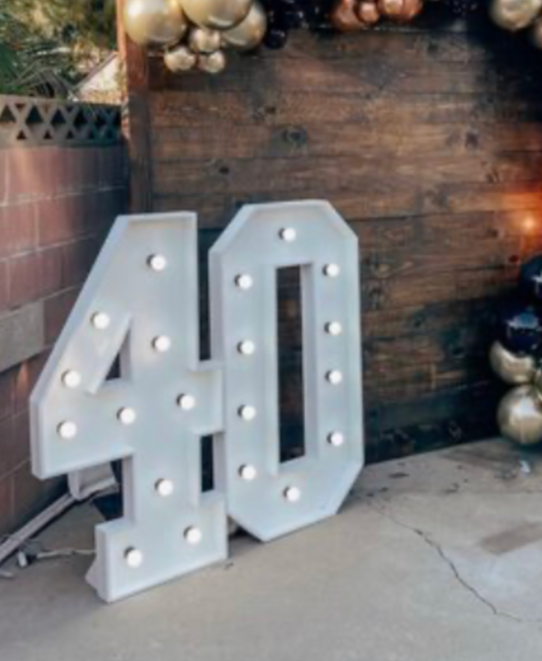 Large white illuminated number 40 sign with bulbs, placed on the ground next to a brick wall and Christmas decorations.
