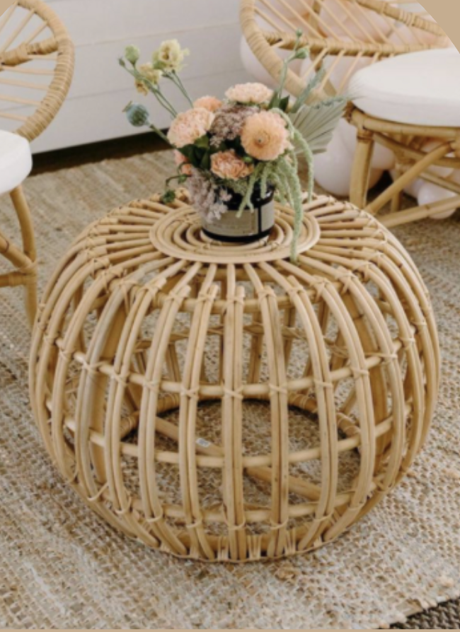 A rattan side table with a round, open design, holding a flower arrangement with various pastel-colored fresh flowers, set on a woven rug in a cozy, modern room.