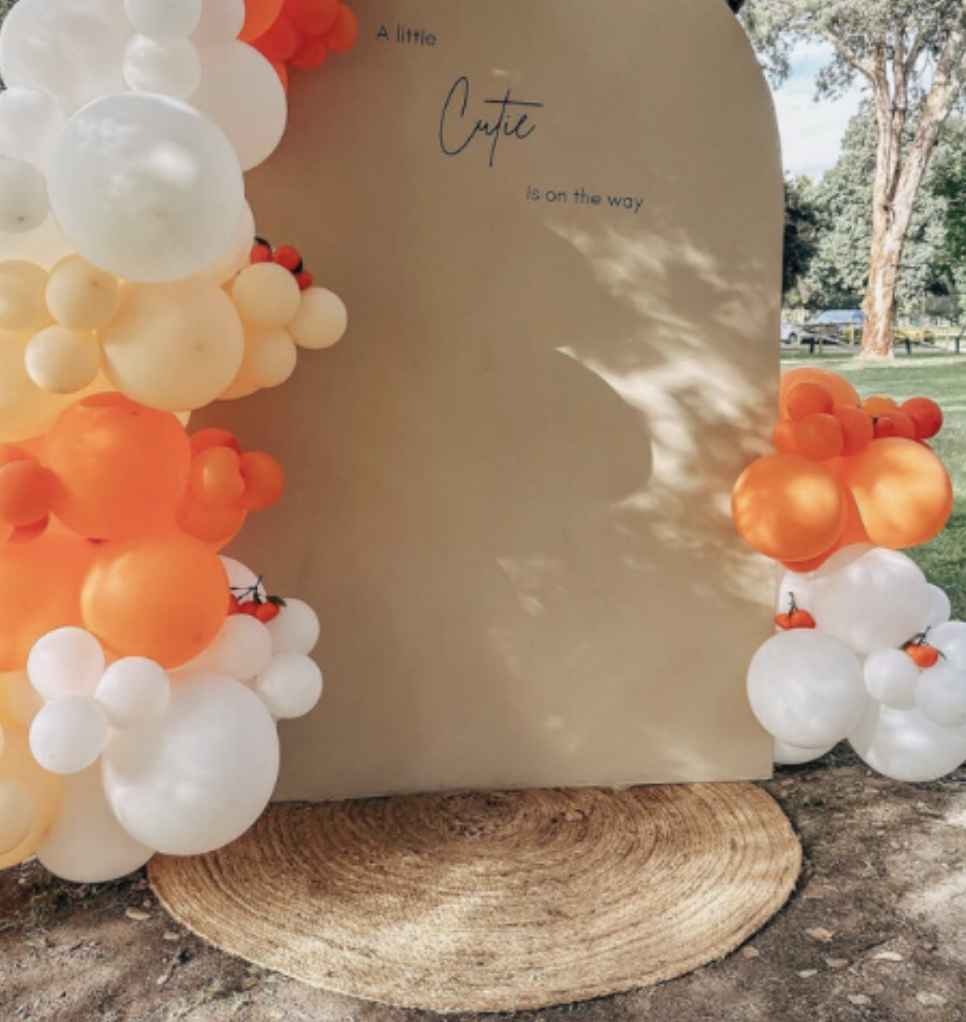 A decorative arch of white, orange, and red balloons surrounding a beige backdrop with the handwritten message 'A little cutie is on the way' for a baby shower or gender reveal event, situated outdoors on a grassy area with trees in the background.