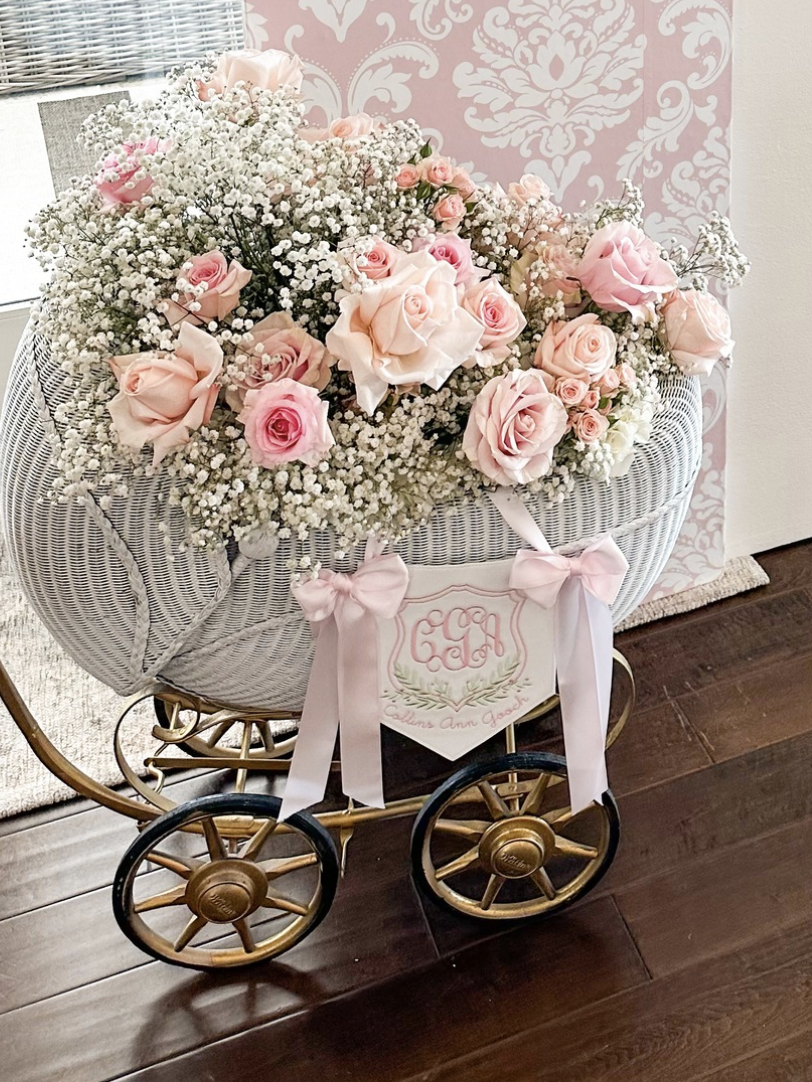 A white wicker baby carriage filled with pink roses and white baby's breath flowers, decorated with pink bows and a monogrammed sign, set on a wooden floor.