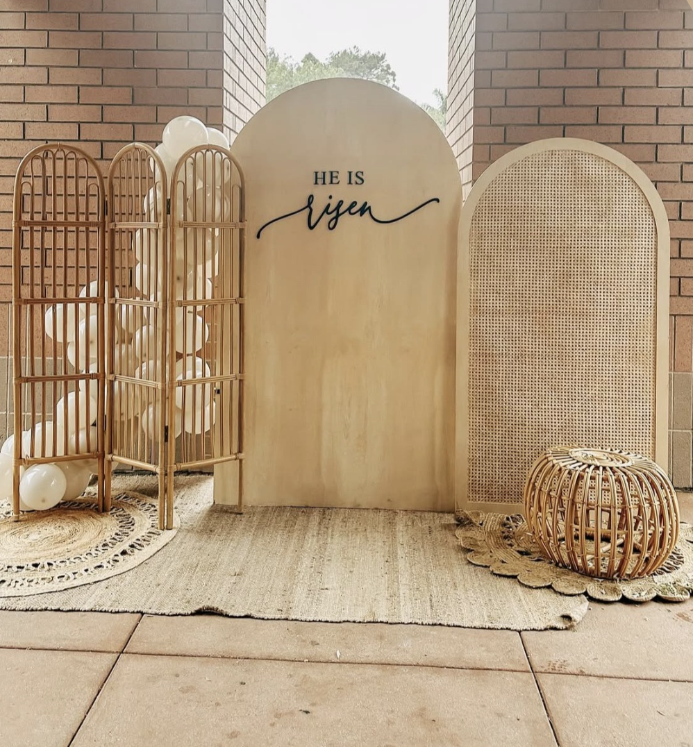 Decorative setup with a three-panel folding screen, a sign that says "He is Risen," and woven furniture on a textured rug, with balloons in the background.
