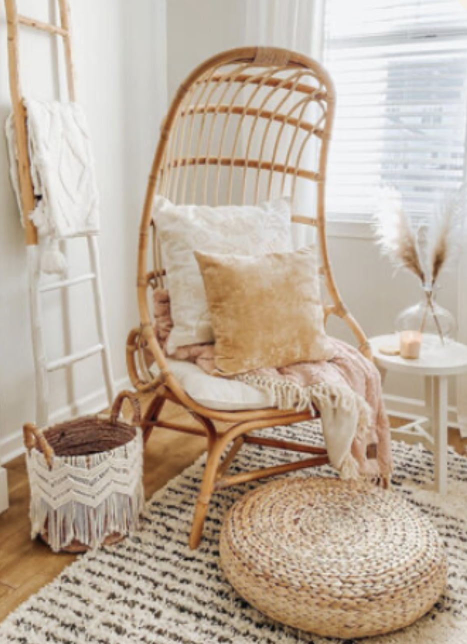 A cozy corner with a rattan high-backed chair adorned with pillows, next to a round rug and a side table with a vase of pampas grass and a candle, in a well-lit room with white walls and blinds.