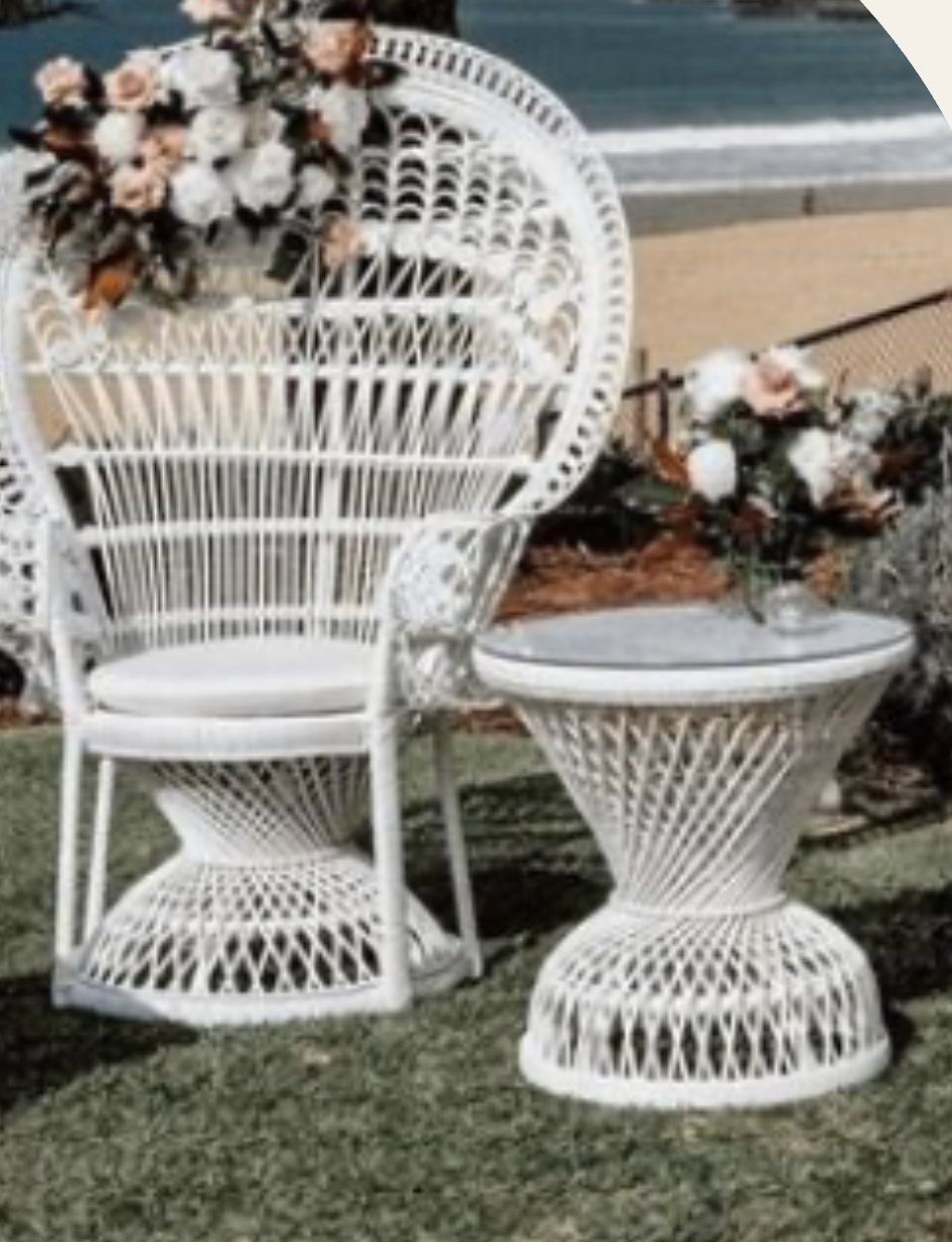 White wicker outdoor chair and side table with floral arrangements, set on grass near a sandy path and a body of water.