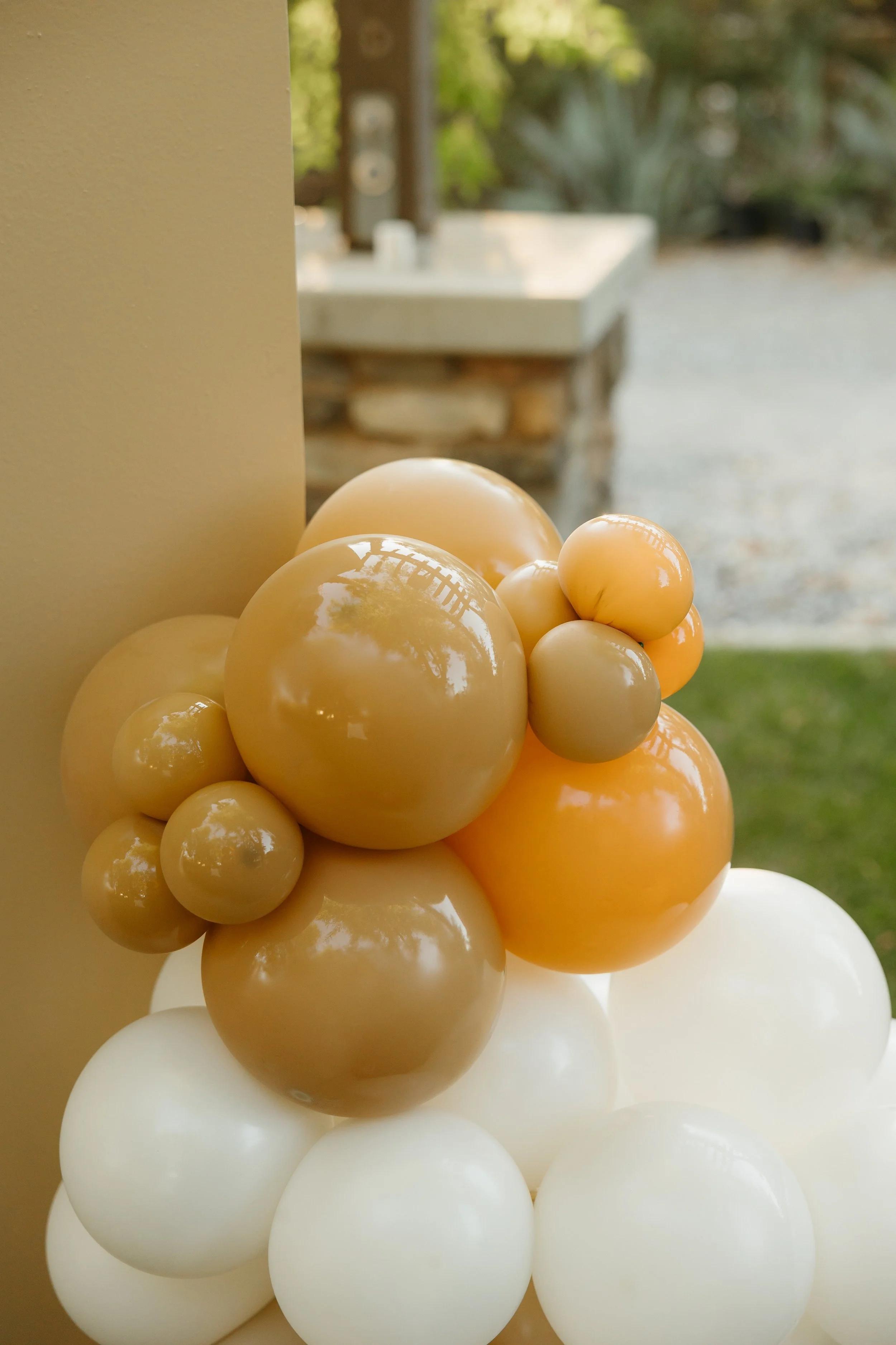 Cluster of cream, beige, and white glossy balloons near a beige wall with outdoor scenery in the background.