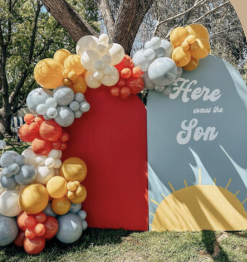 Colorful balloon arch with white, yellow, orange, and red balloons surrounding a red and blue backdrop with the words "Here comes the Sun" and a sun illustration.