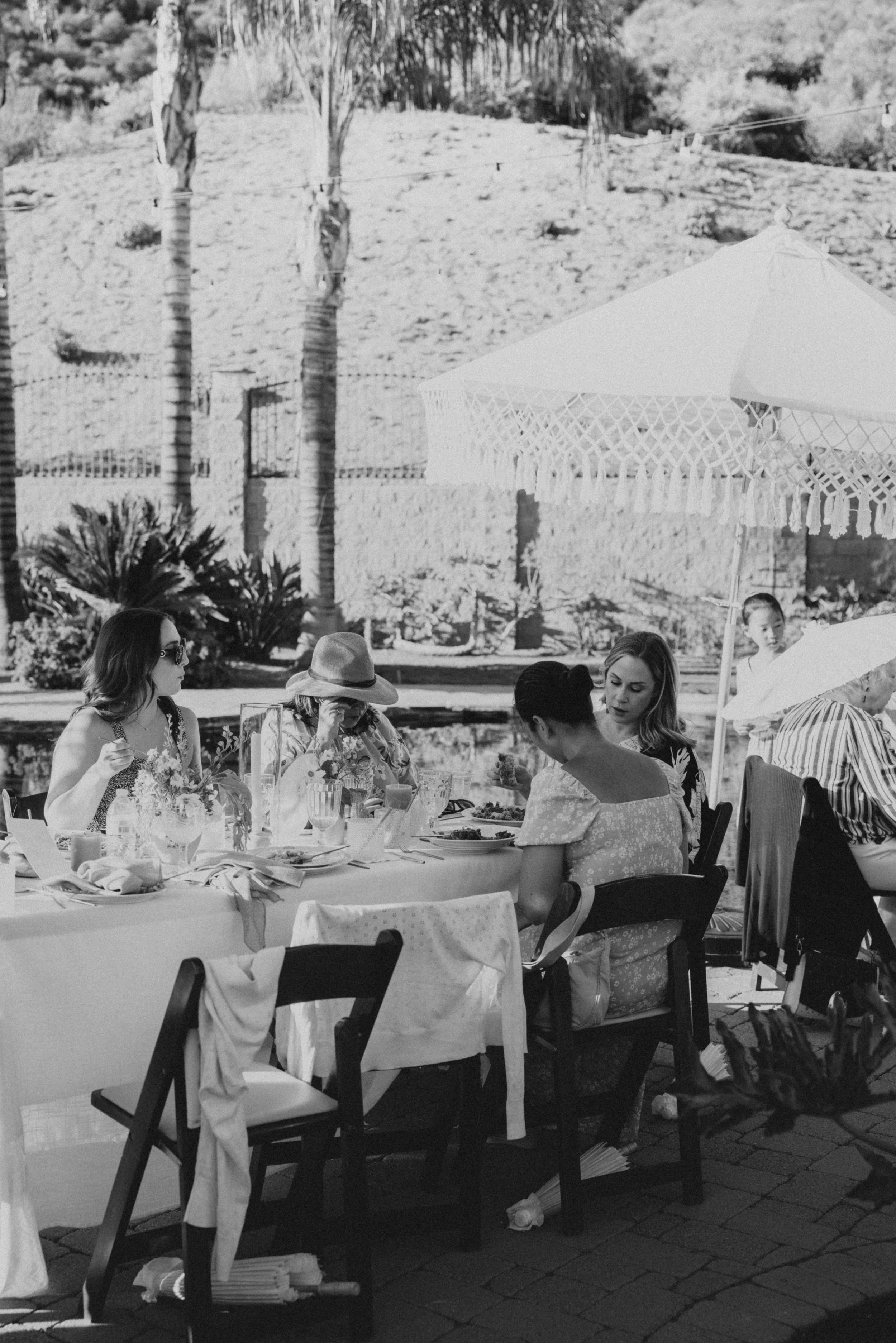 People sitting around a table outdoors, enjoying a meal under large umbrellas, with trees and a hillside in the background.