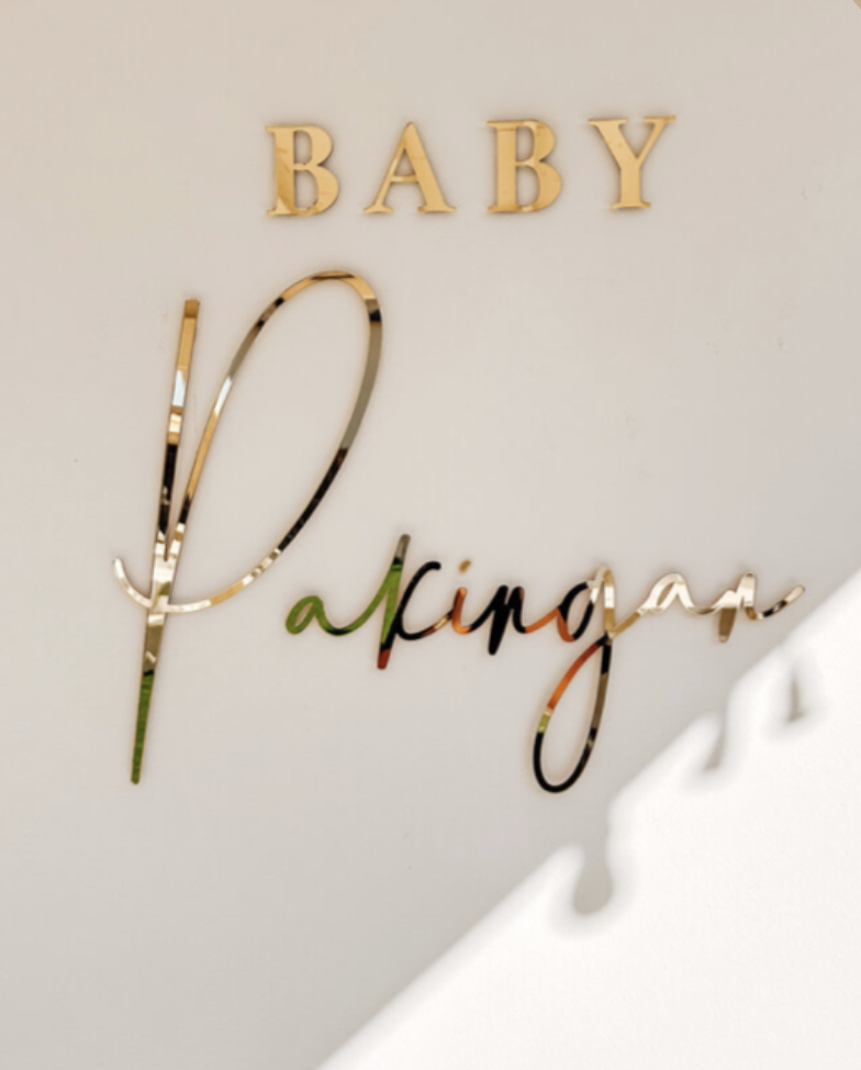 Decorative wall sign saying 'Baby Pacifier' with the words 'Baby' in wooden letters and 'Pacifier' in metallic, cursive script on a white wall.