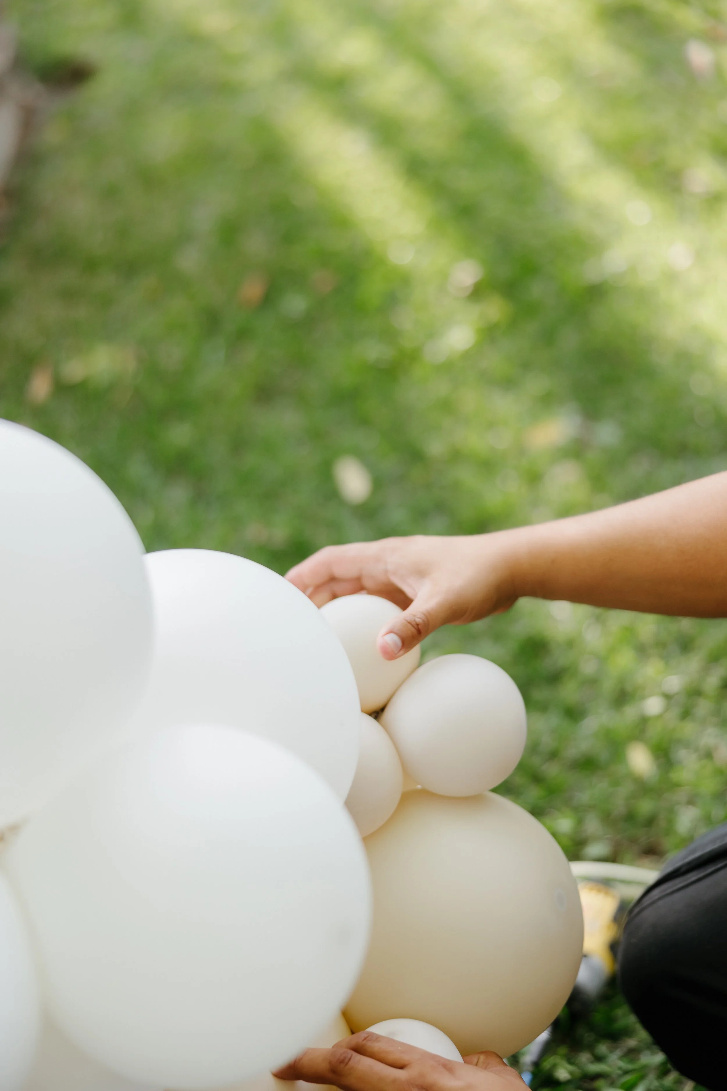 Person arranging white helium balloons outdoors on green grass.