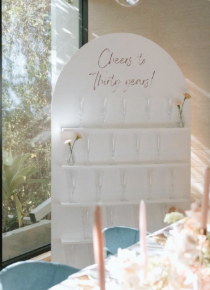 A white standing display with the message 'Cheers to Thirty years!' written on it, decorated with small flowers in glass vases, and set in a bright room with large windows and greenery outside.