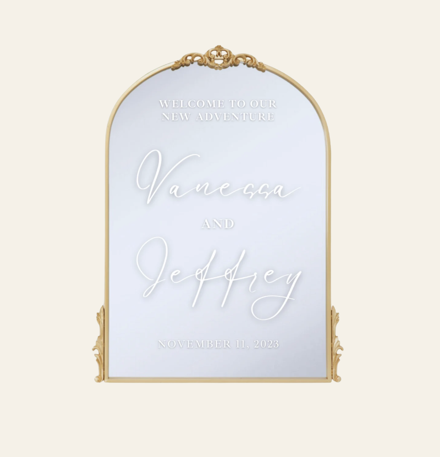 A decorative wedding mirror with gold frame, engraved with the message 'Welcome to our new adventure Vanessa and Jeffrey, November 11, 2023'.