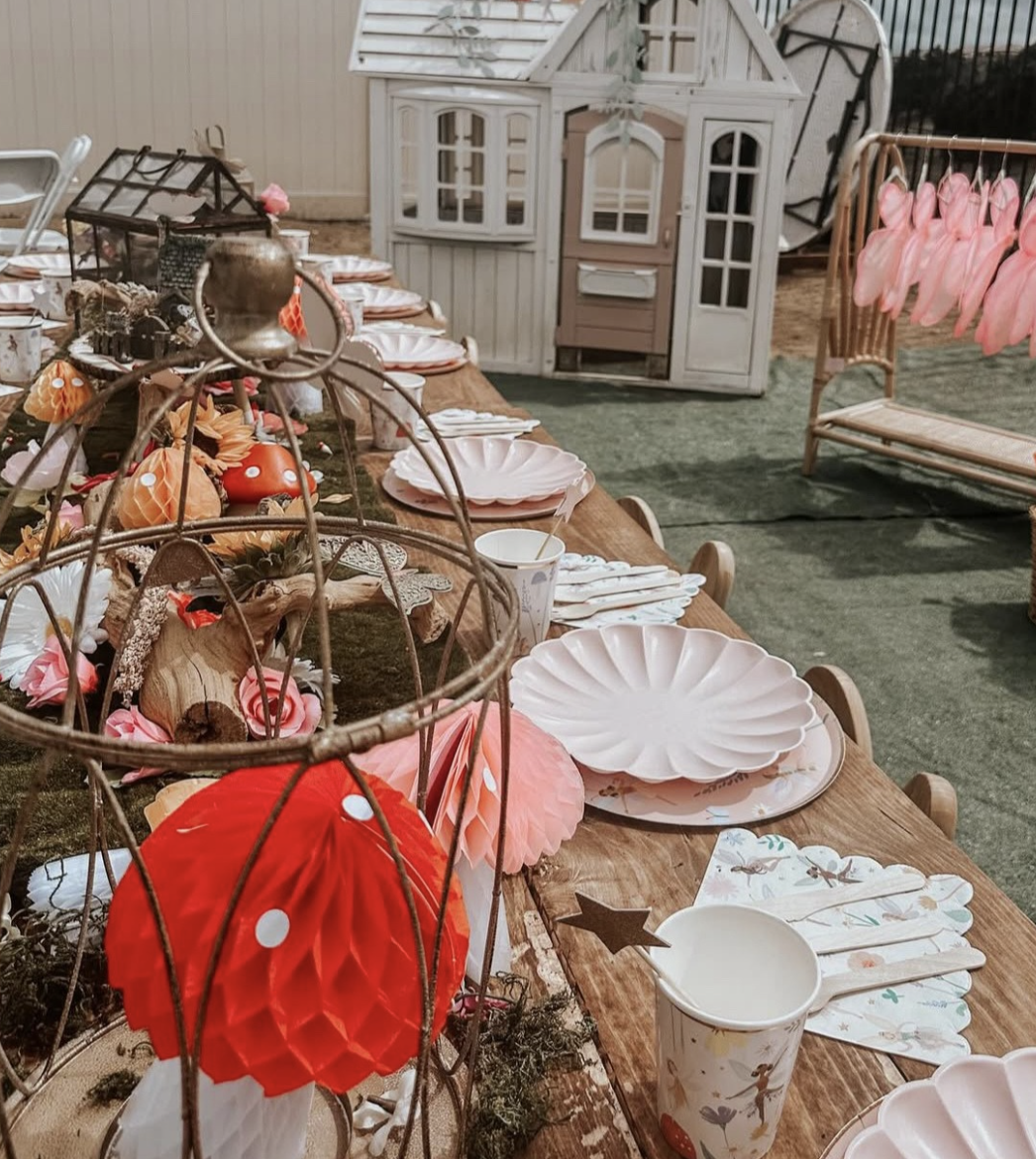 A decorated outdoor party table with pink and white plates, paper napkins, and cups featuring a duck design. The table is adorned with floral and mushroom-themed decorations, including a red honeycomb paper lantern and a birdcage centerpiece. In the background, there is a small wooden playhouse and a pink bench with pink clothing hanging on a rack.