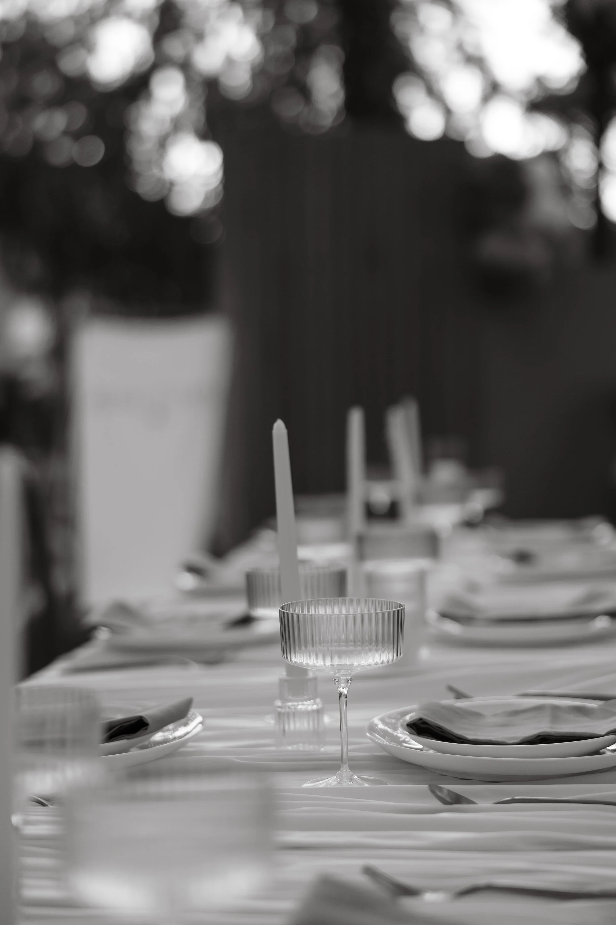 A table set for a meal with plates, silverware, candles, and glasses, outdoors with blurred background.