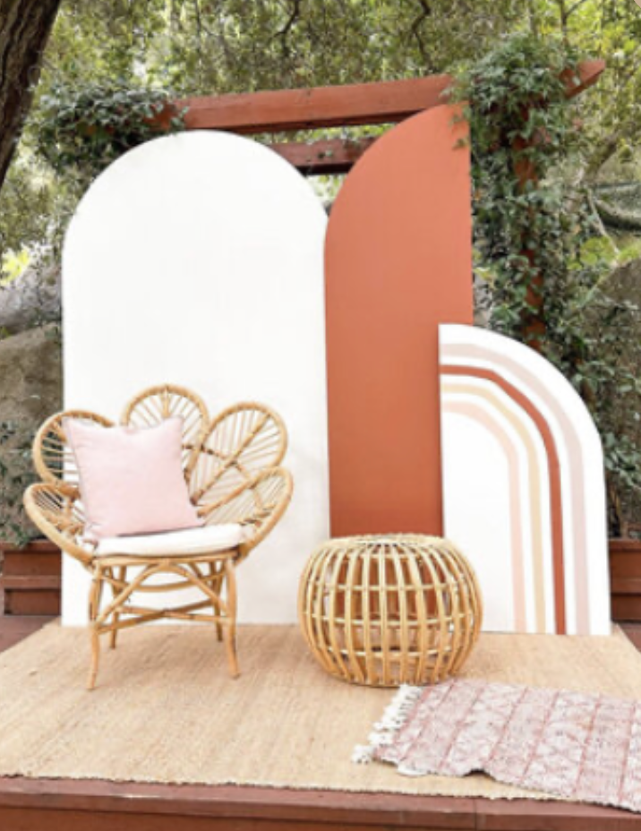 Outdoor decorative backdrop with urban boho theme, featuring painted geometric shapes in white and terracotta, rattan chair with pink pillow, round wicker table, and a pink patterned rug on a wooden deck surrounded by greenery.