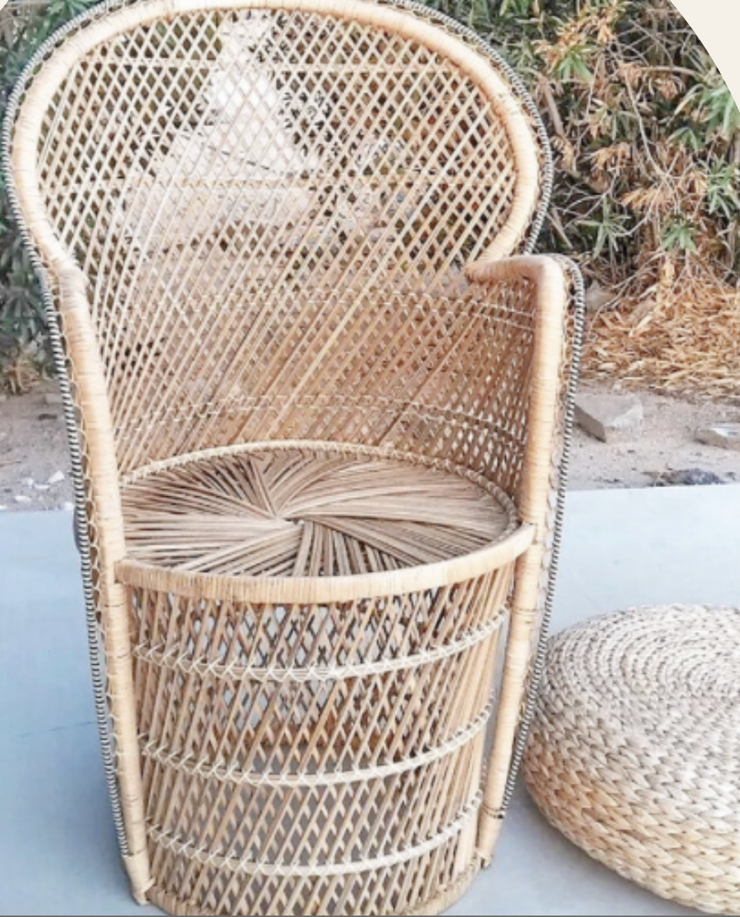 A tall rattan chair with a round woven bottom, curved backrest, and armrests, placed outdoors on concrete with plants and rocks in the background, next to a round woven ottoman.