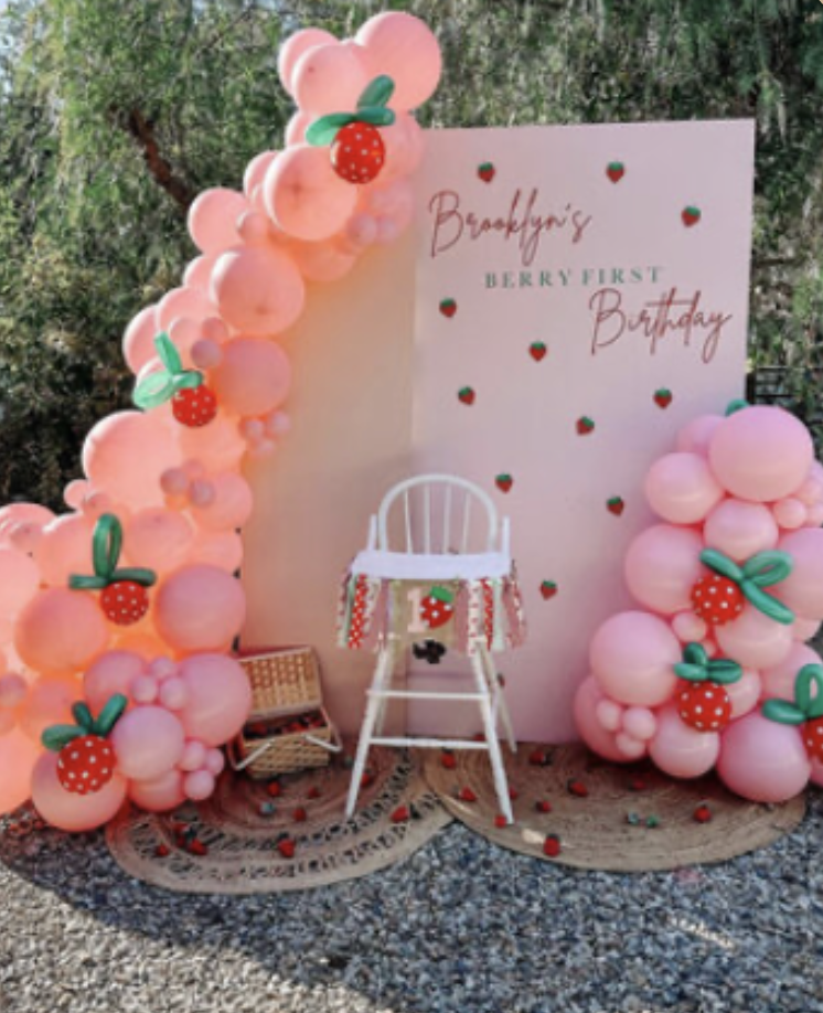 Decorative setup with a pink balloon arch decorated with strawberry cutouts, a pink backdrop with the text "Brooklyn's Berry First Birthday" and small strawberry decals, a white chair with a strawberry-themed tablecloth, and a small wicker basket on a light-colored rug outdoors.