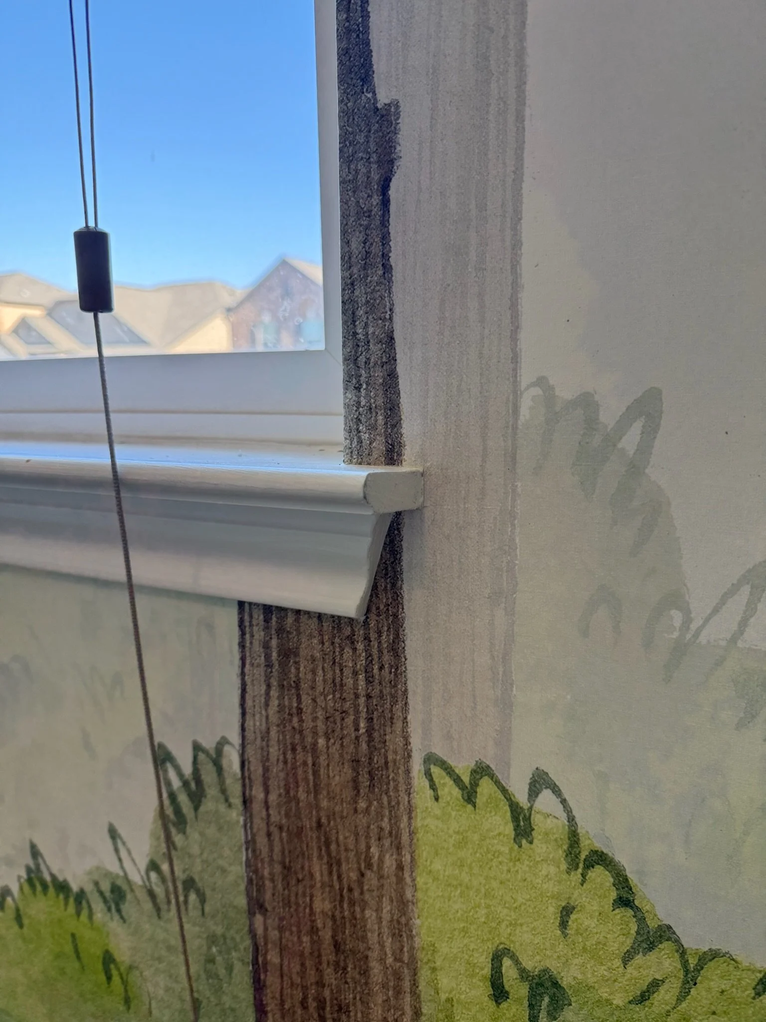 Close-up view of a window with a white window sill, partially painted wall, and a view of houses outside on a sunny day.