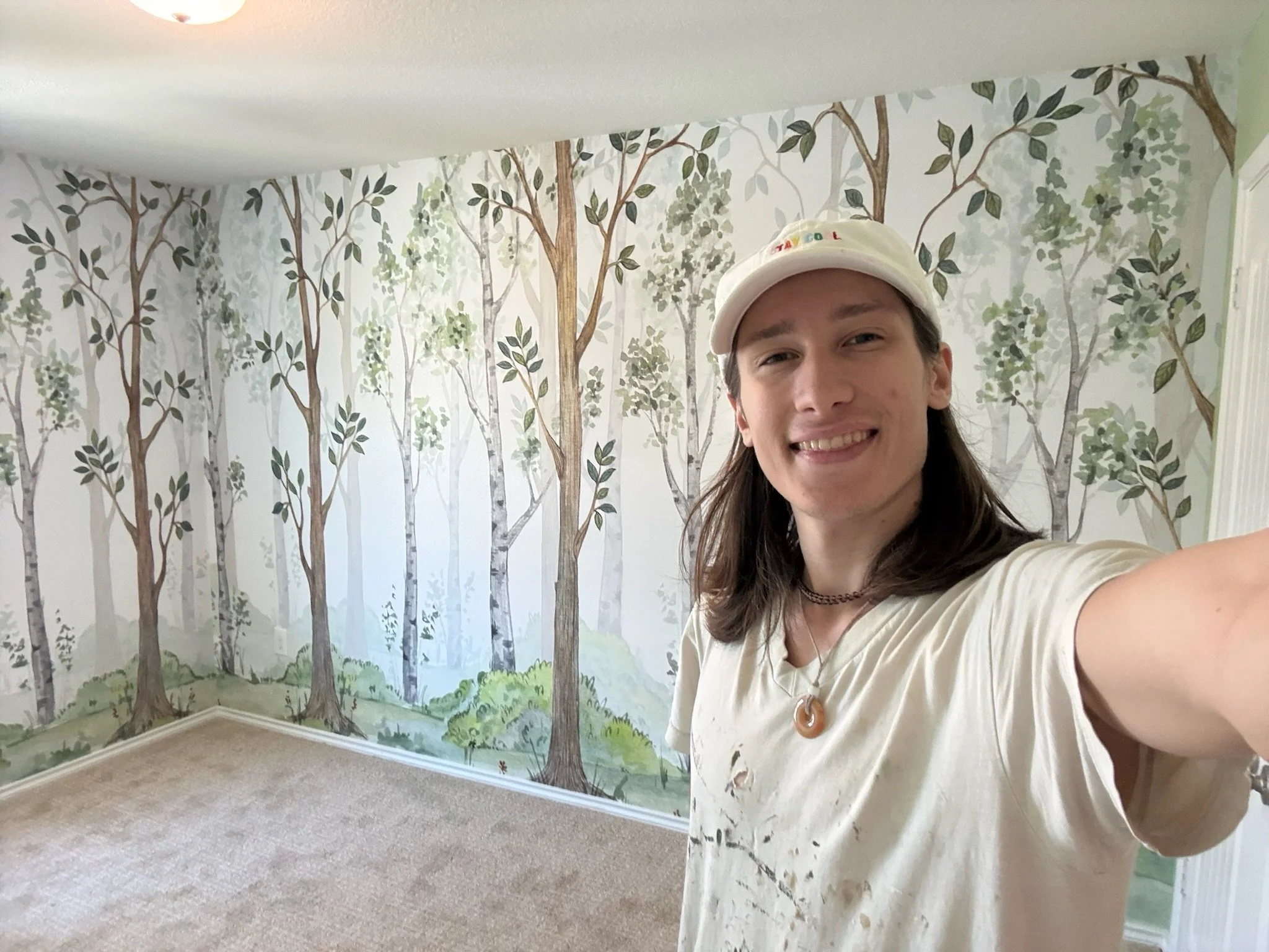 A person taking a selfie in a room with forest-themed wall mural featuring trees and greenery.