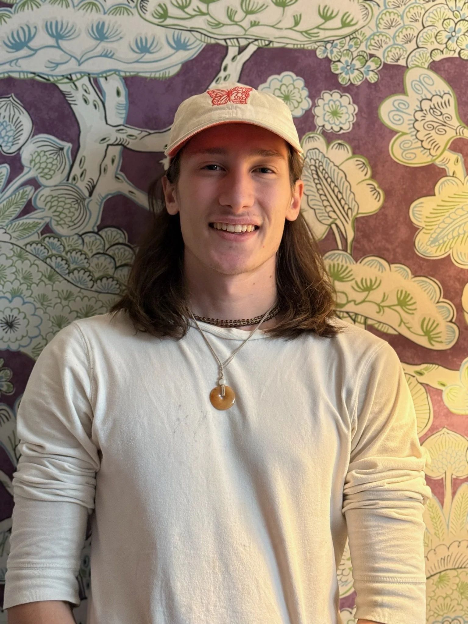 Young man with long hair wearing a beige cap and a cream-colored long-sleeve shirt, standing in front of a colorful botanical mural.