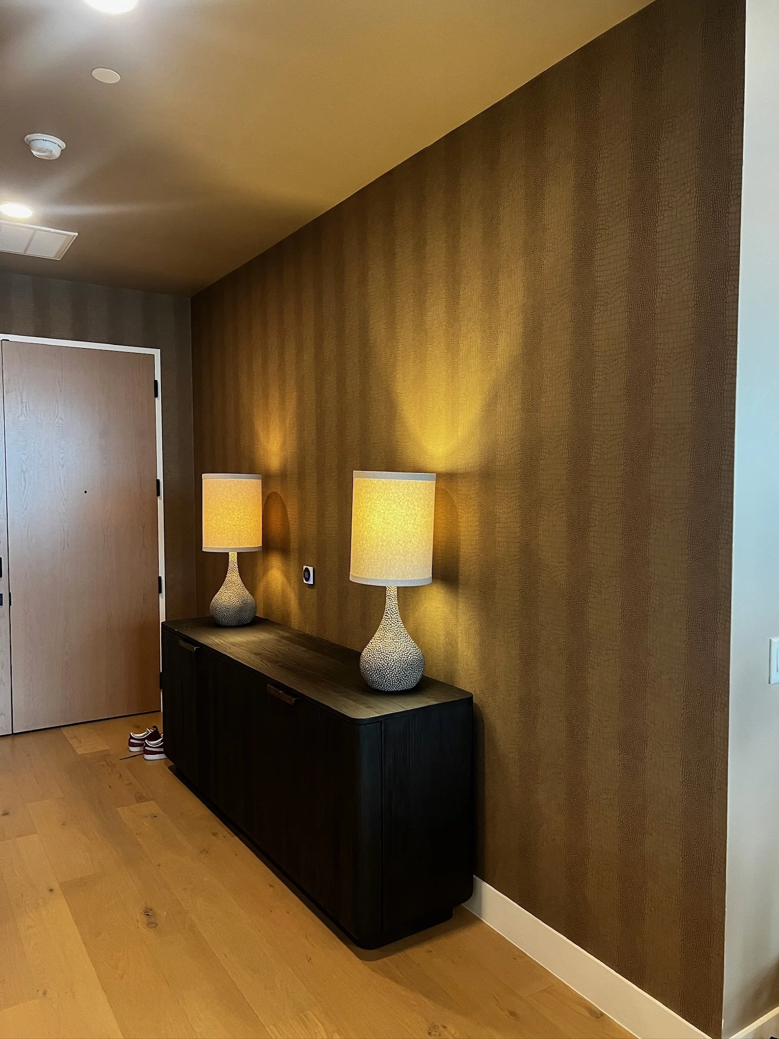 A wooden console table with two decorative table lamps with yellow lampshades on top, set against a textured brown wall in a modern interior space.