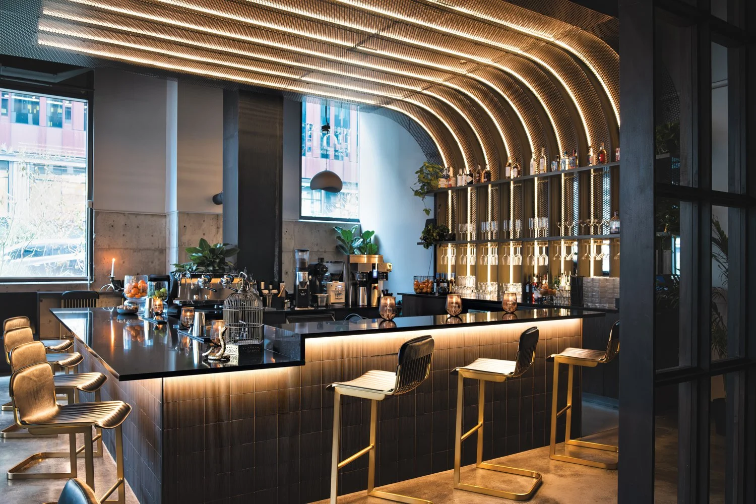 Modern bar with black countertop, gold barstools, illuminated arch decor, shelves with bottles, windows with city view, interior plants, and ambient lighting.