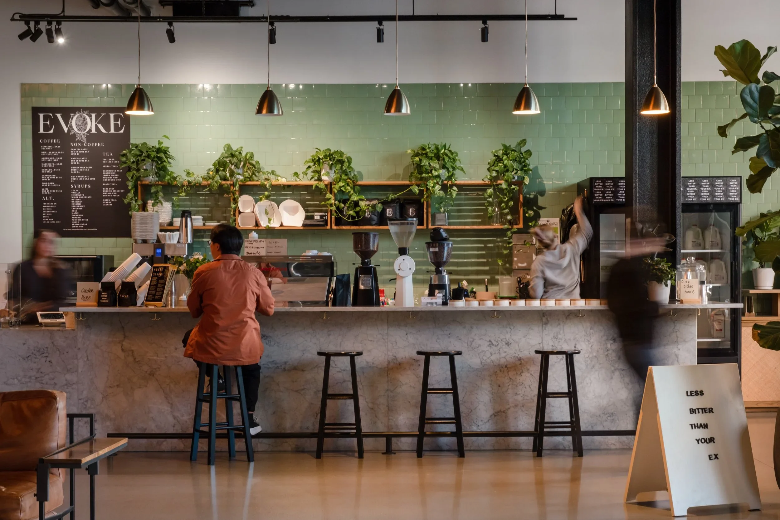 Inside a modern cafe or coffee shop with a marble counter and bar stools. Behind the counter are shelves with potted plants and a menu board on the wall. There are people ordering and working behind the counter, with warm lighting and green tiled wall.