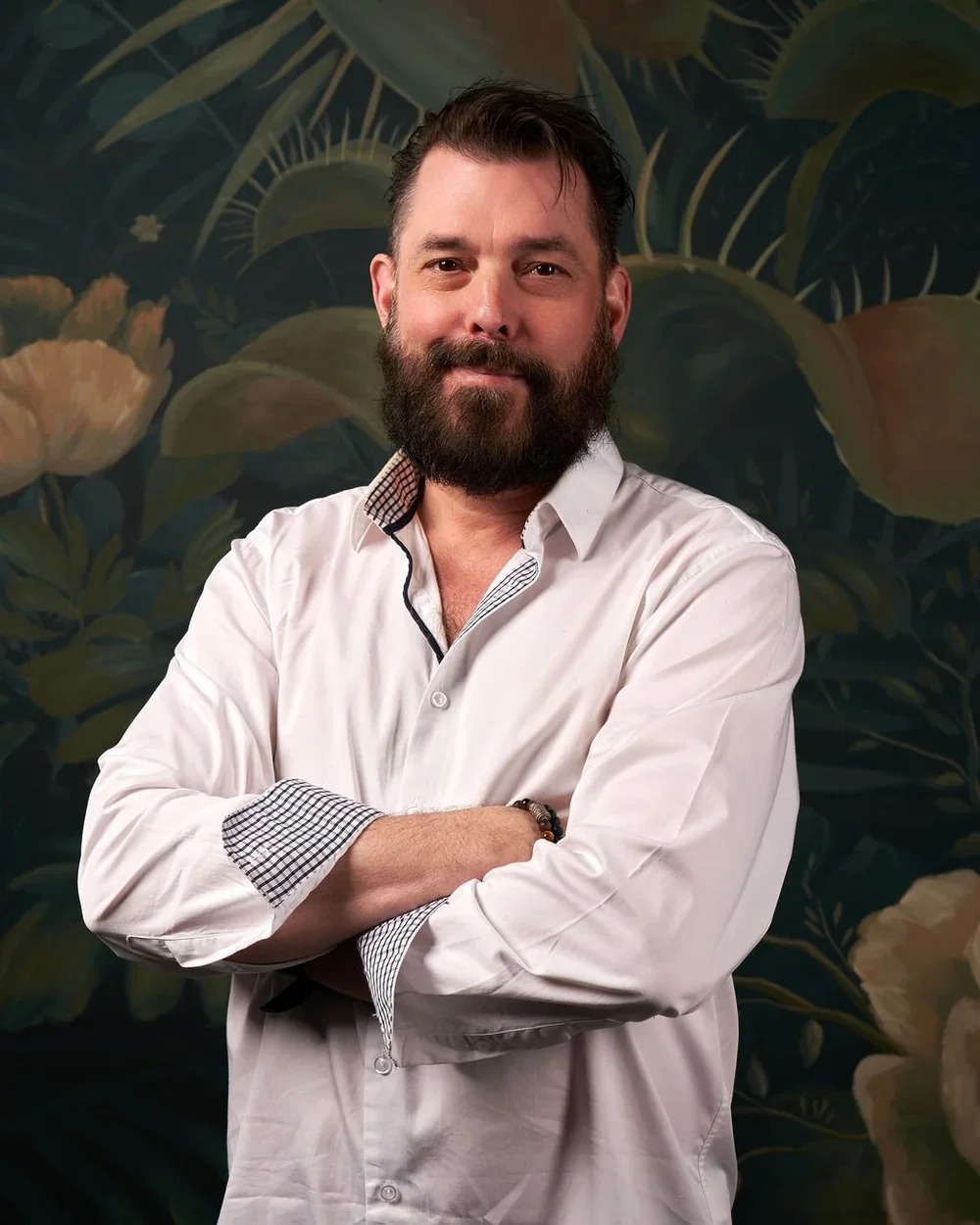 A man with a beard and mustache, wearing a white dress shirt with checkered cuffs, standing with arms crossed in front of a dark leafy background.