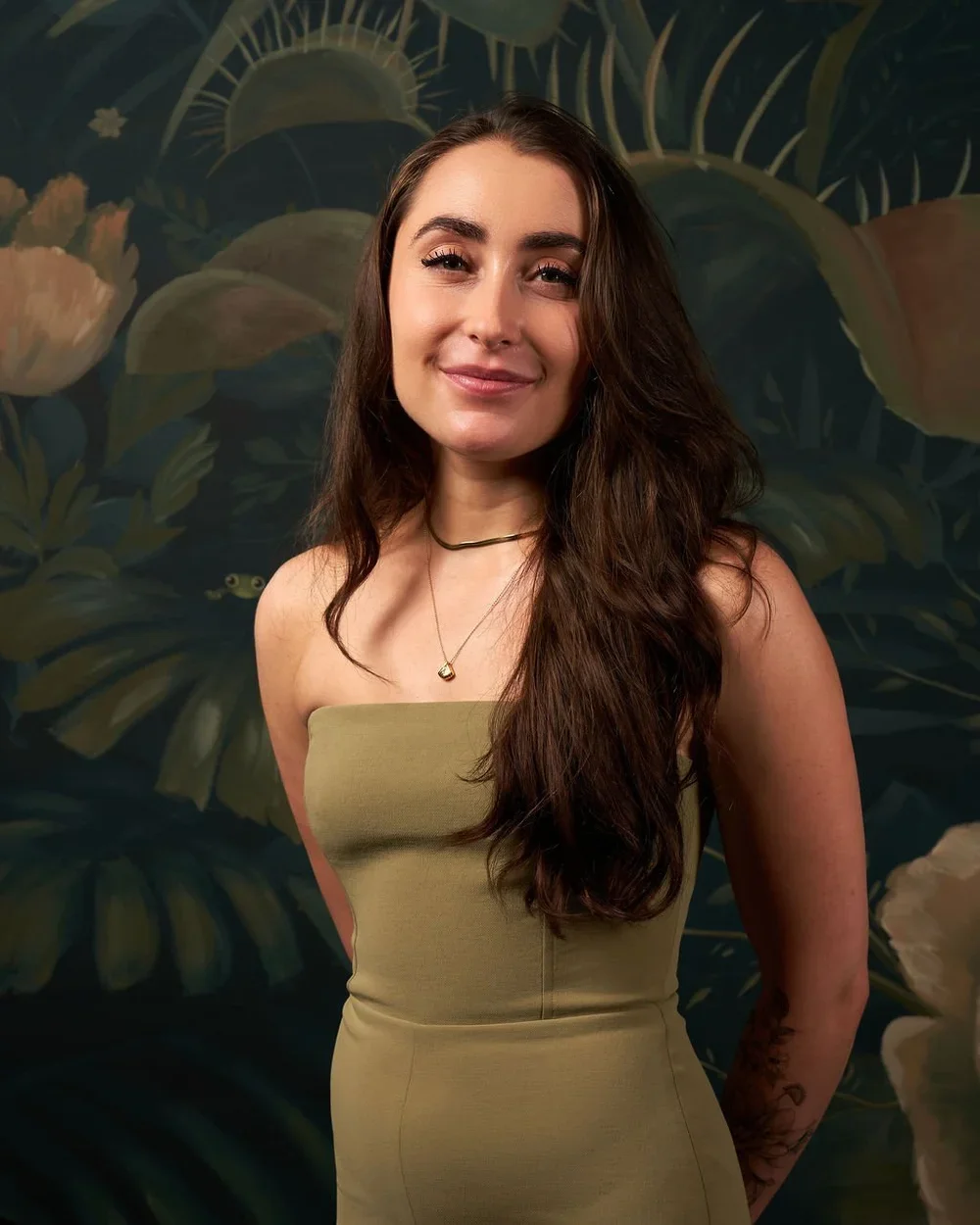 A woman with long, wavy brown hair and light skin, smiling, wearing a strapless olive green dress with a necklace and a choker, standing in front of a dark green floral mural background.