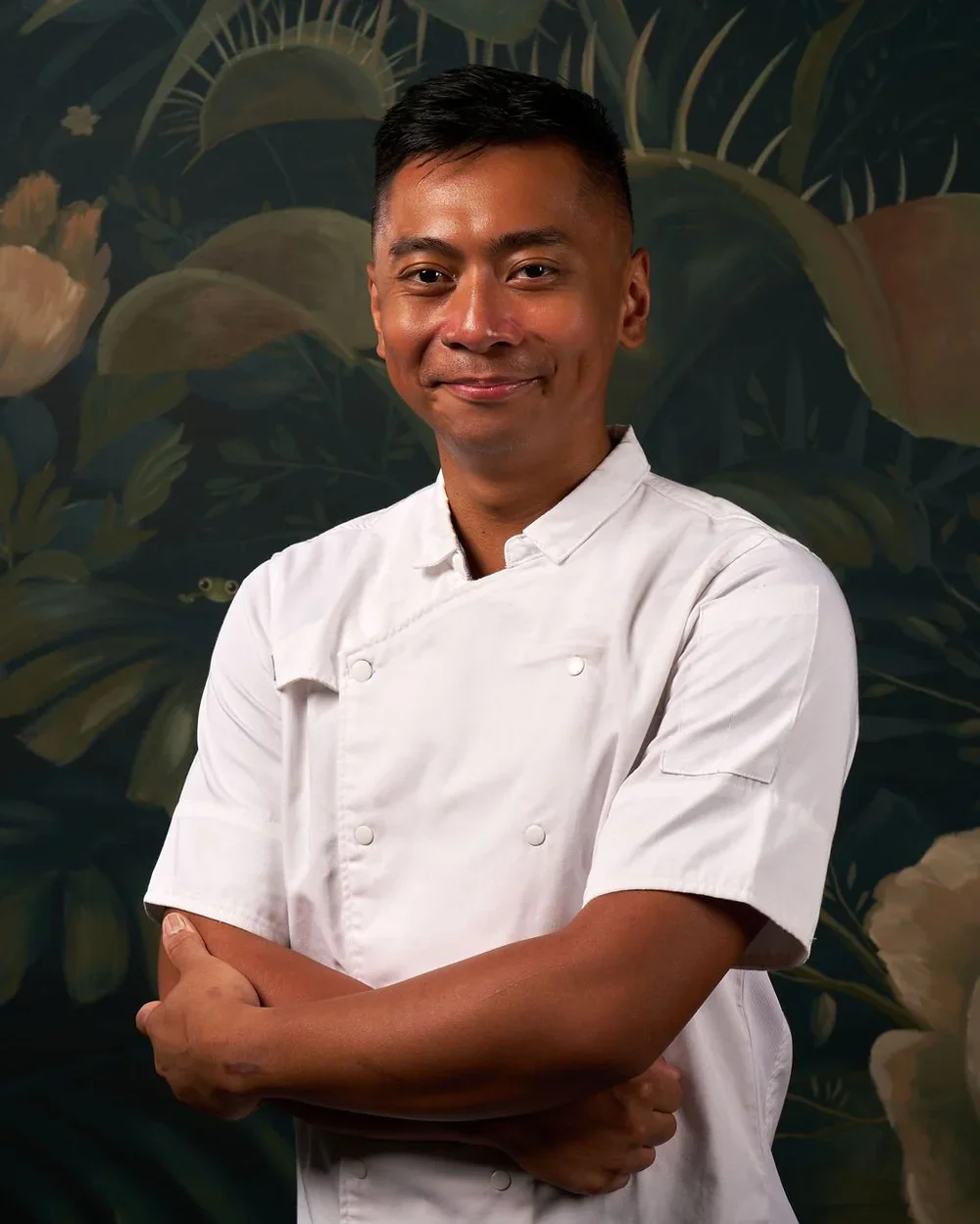 A smiling chef in a white uniform with arms crossed standing in front of a dark, botanical background.