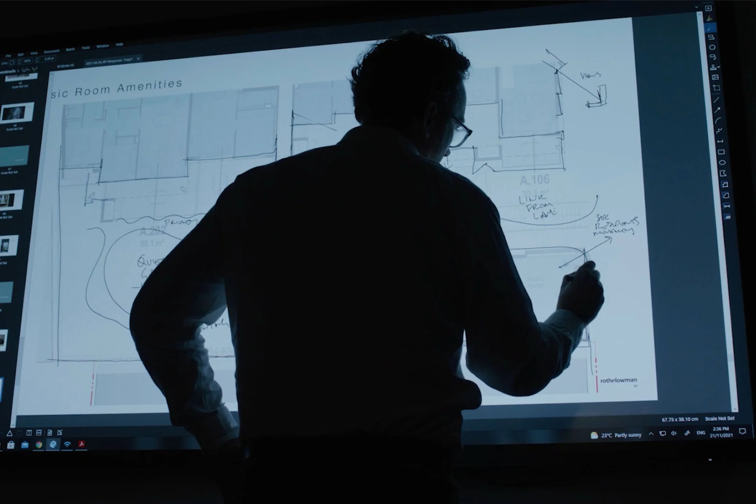 A person with glasses is standing in front of a large digital screen, drawing on a floor plan titled 'Room Amenities' with a stylus. The screen displays architectural sketches and notes.