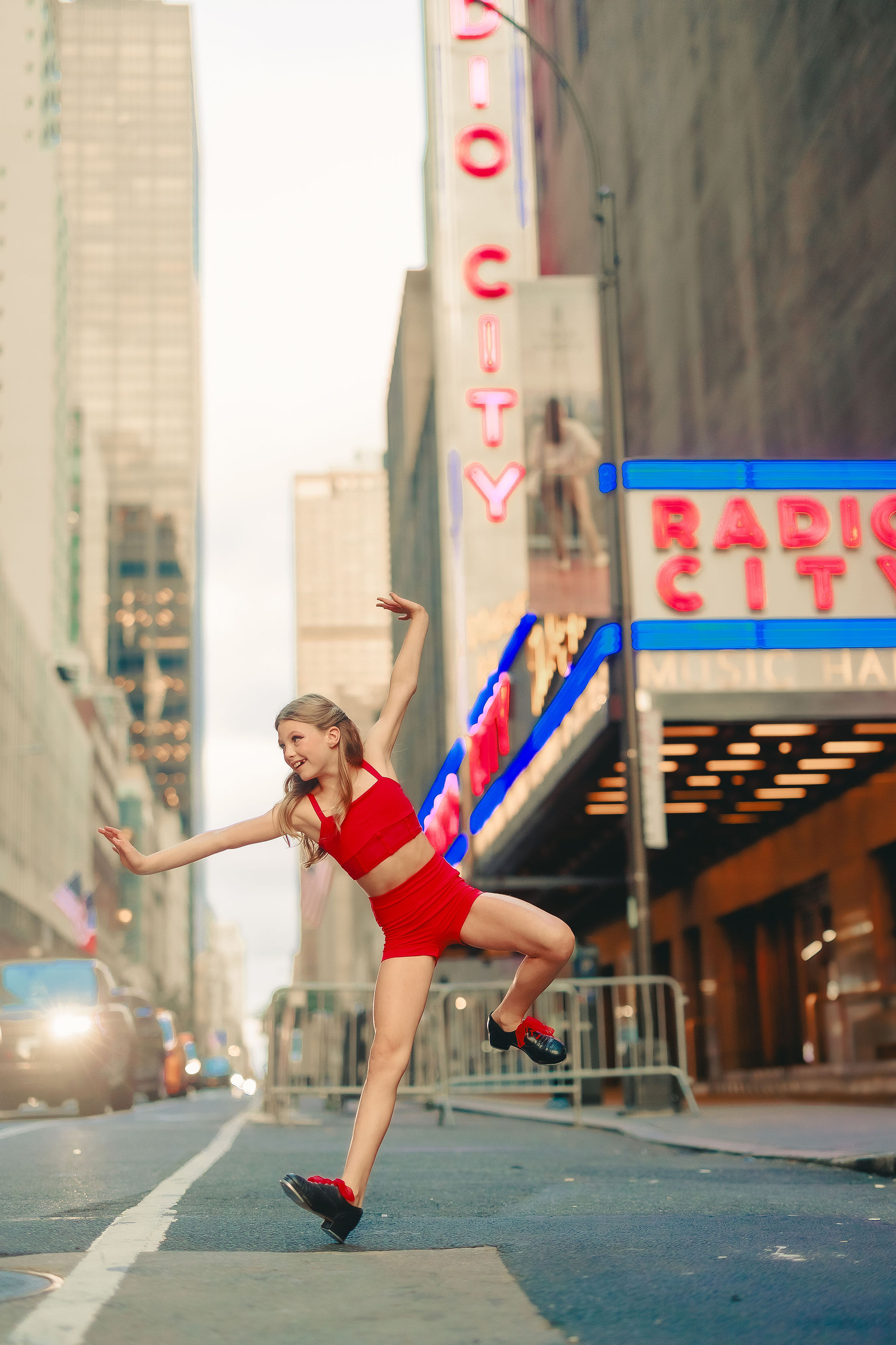 A young woman in a red outfit dancing or jumping on a city street at dusk, with neon signs and tall buildings in the background.