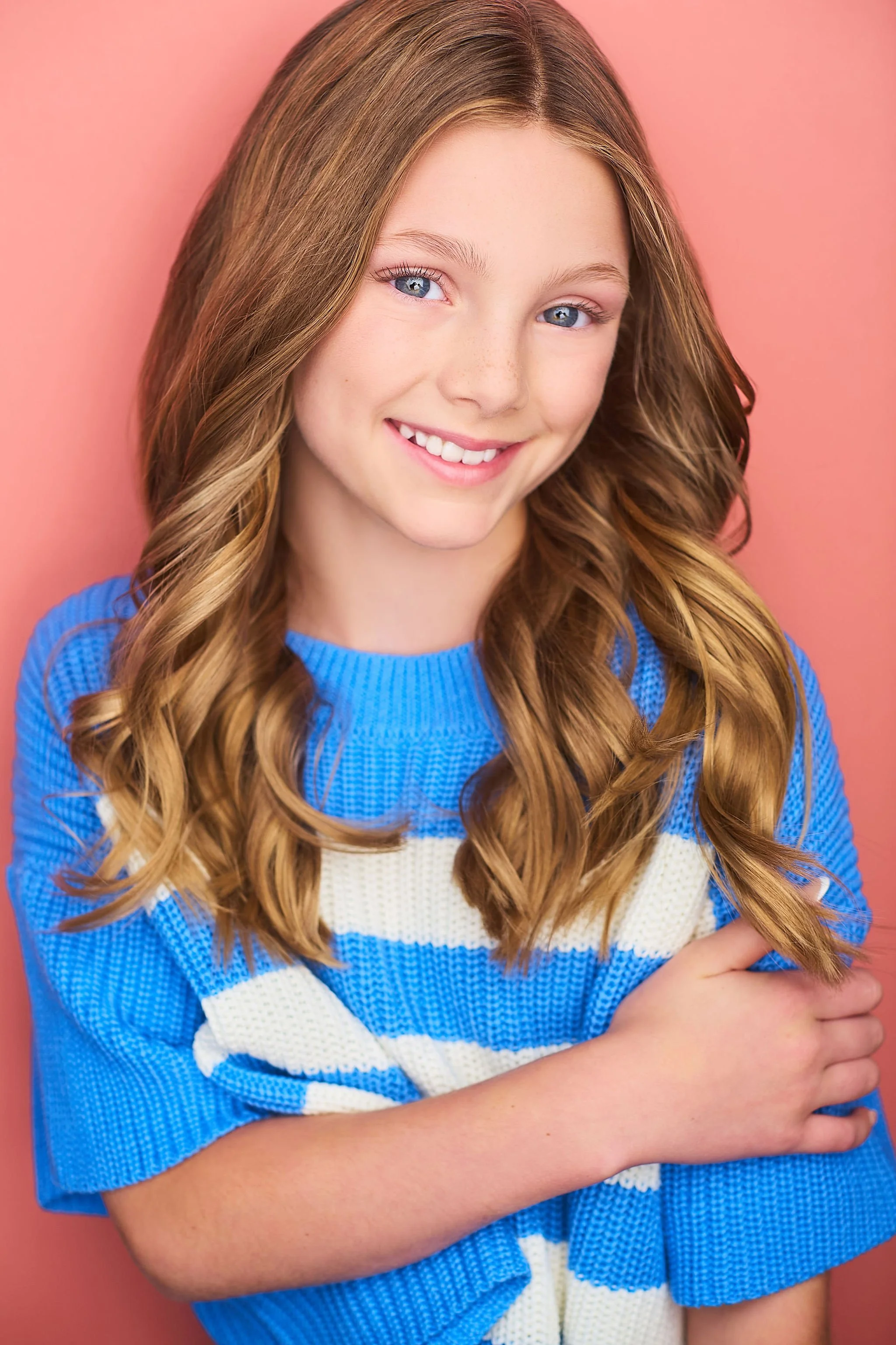 A young girl with long wavy red hair, blue eyes, and freckles, smiling and wearing a blue and white striped shirt, against a pink background.