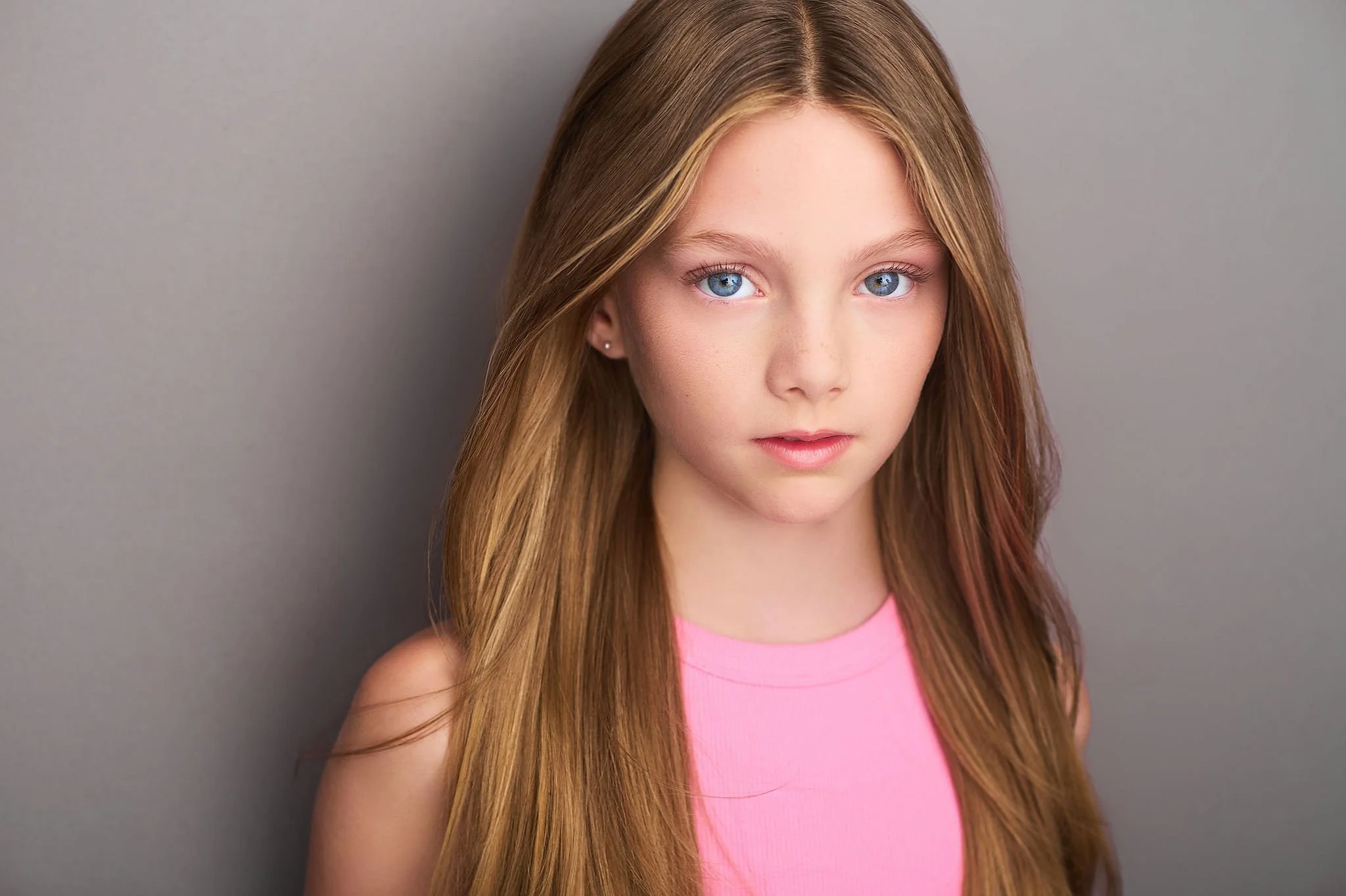 A young girl with long, light brown hair and blue eyes, wearing a pink sleeveless top, standing against a plain gray background.