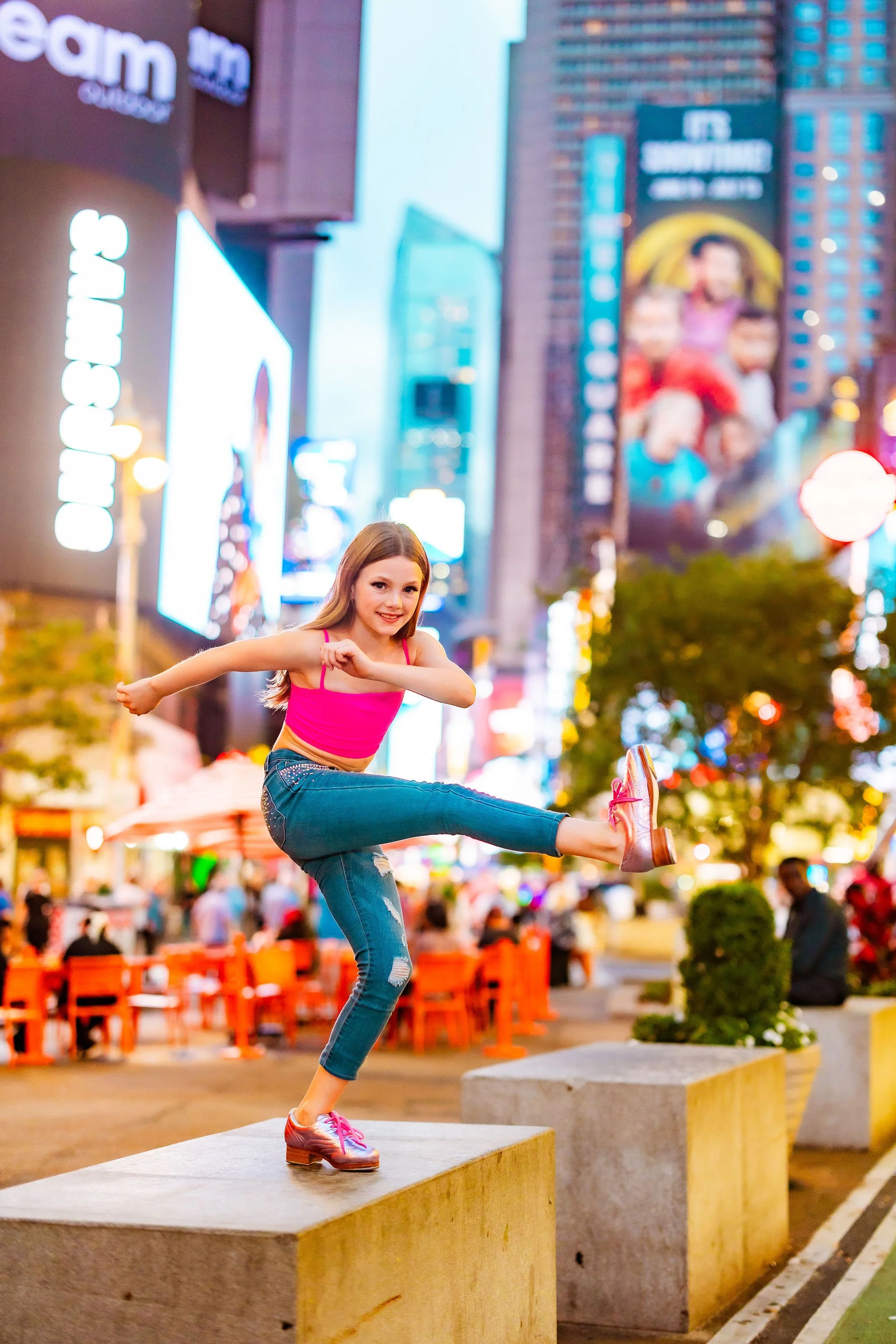 A young girl in a pink top and jeans performing a high kick on concrete blocks in an urban area at night, with illuminated billboards and crowded outdoor seating in the background.