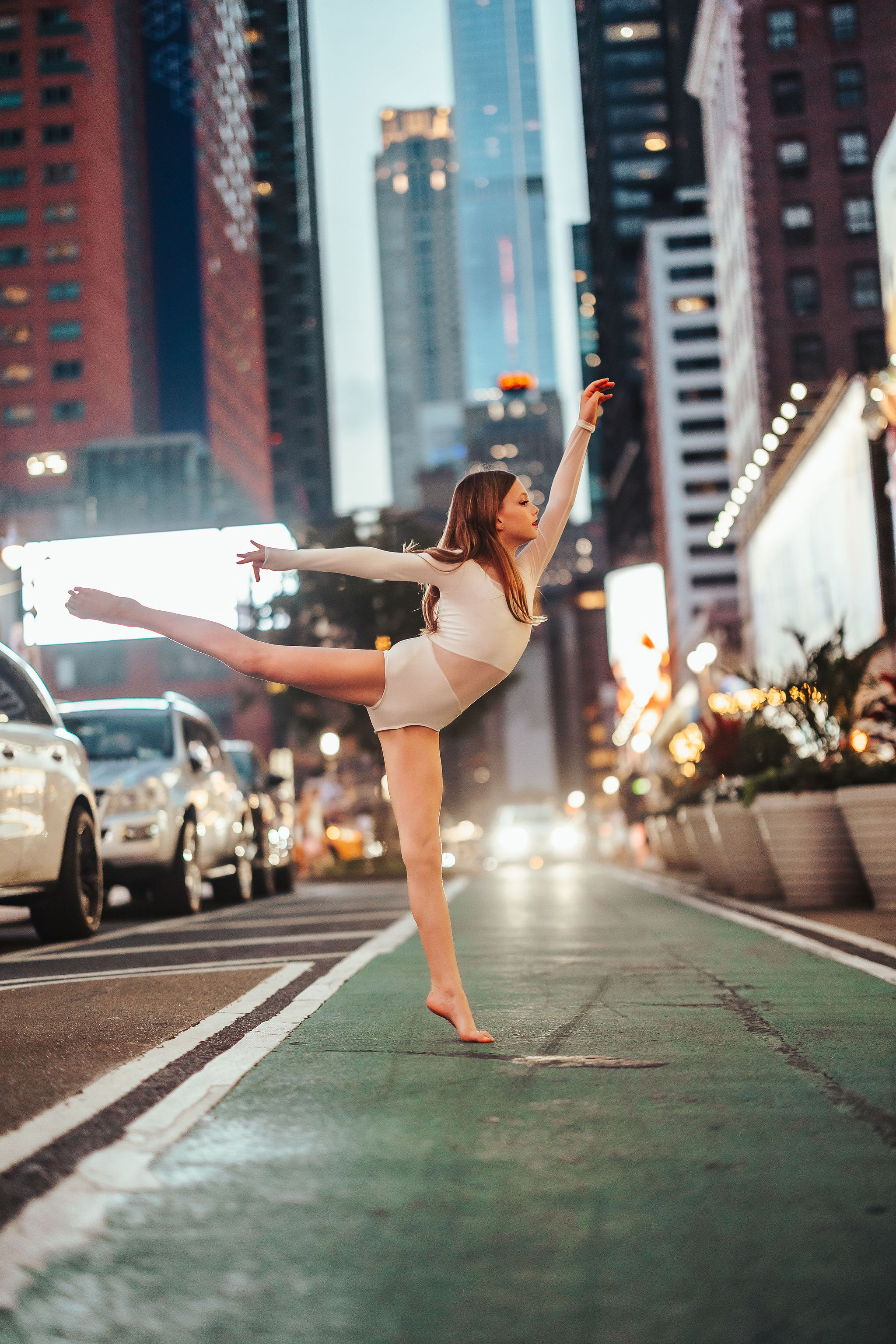 A woman in a white dance outfit performs a ballet pose on a city street with tall buildings and cars in the background.