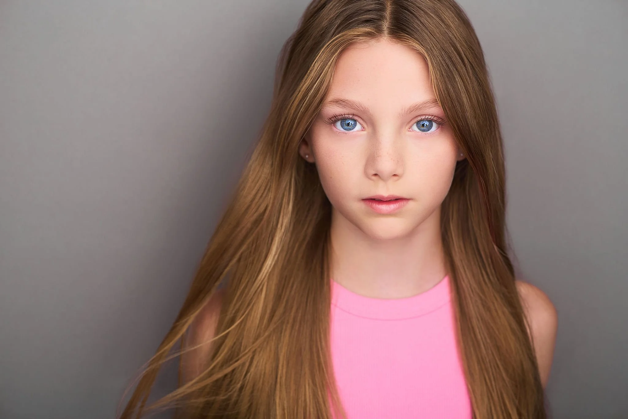 A young girl with long, straight, red hair and blue eyes wearing a pink sleeveless top, standing against a plain gray background.