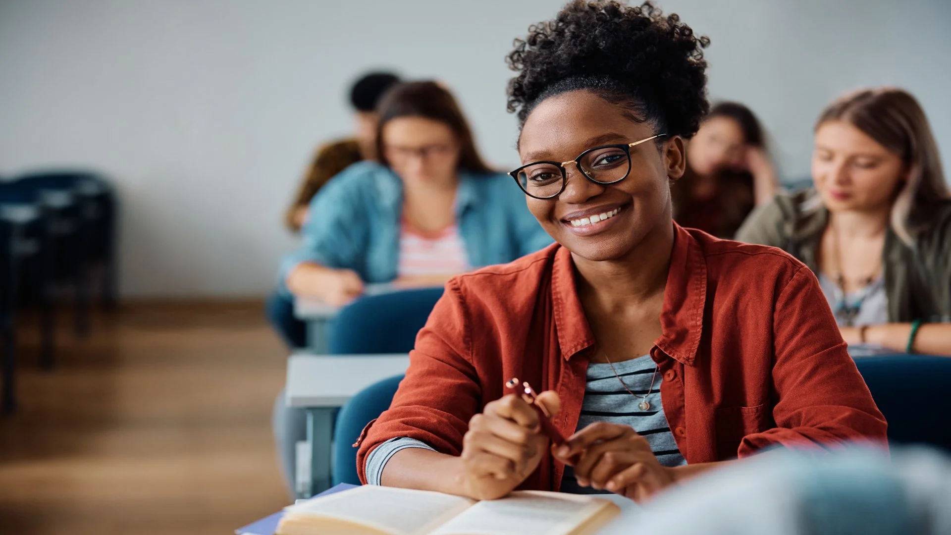 A smiling young woman with glasses and curly hair, sitting in a classroom with open books in front of her, and other students in the background.
