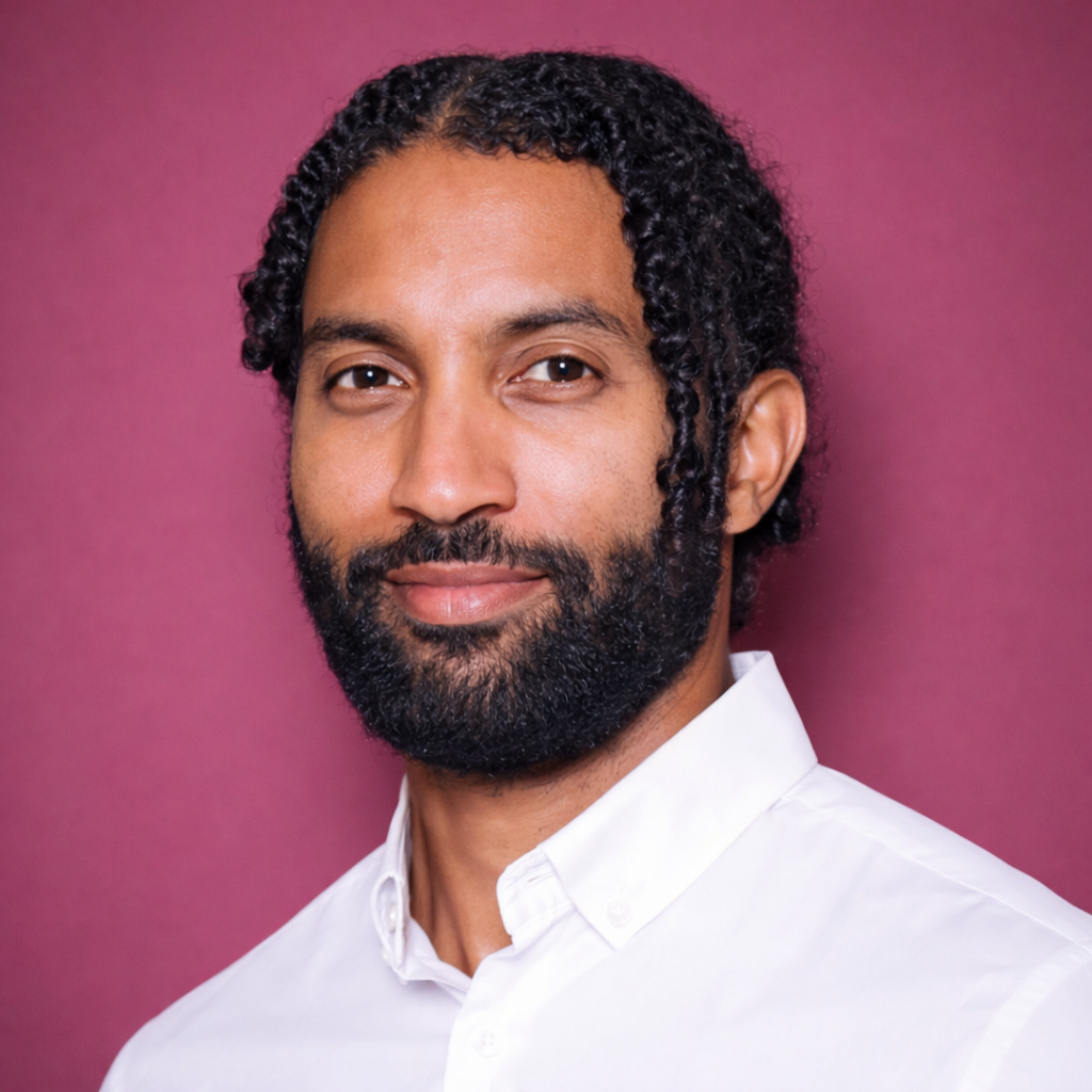 Close-up of a man with dark curly hair and a beard, wearing a white button-up shirt, standing against a pink background.