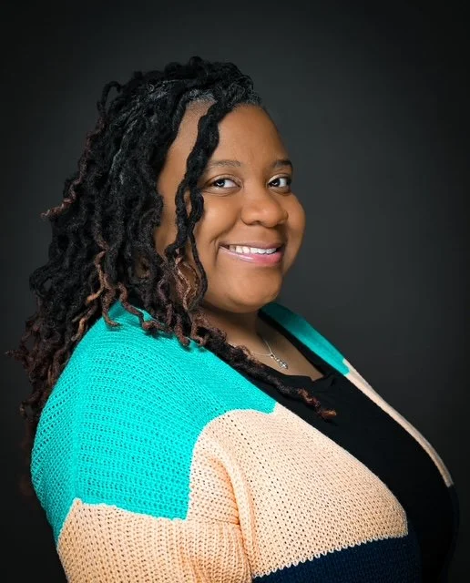 Portrait of a smiling Black woman with curly hair, wearing a multicolored cardigan and black top, against a dark background.