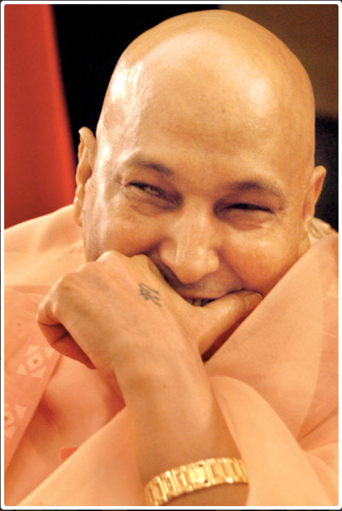Guruji Maharaj smiling and covering his mouth with his hand, wearing a light-colored garment and a gold watch.
