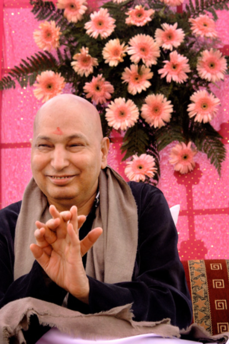 Guruji Maharaj with a slight smile, sitting in front of a pink floral backdrop, wearing traditional attire and making a gesture with his hands.