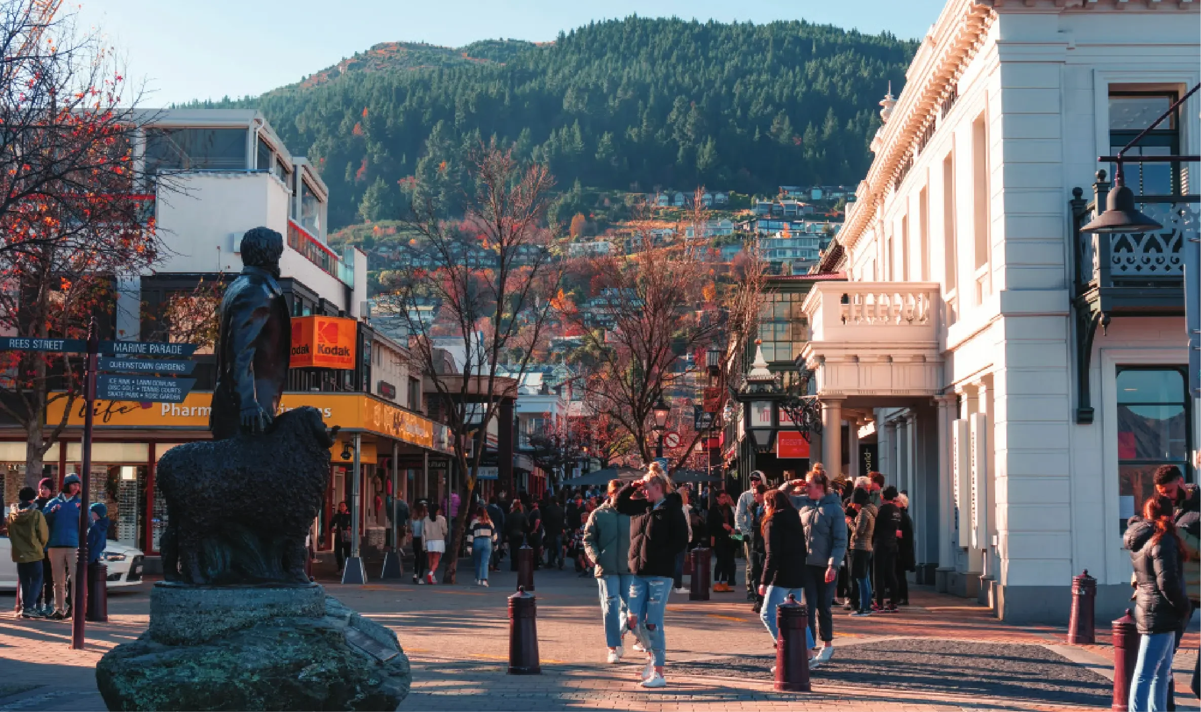 Street scene in a small town with people walking, colorful storefronts, trees with autumn leaves, a statue of a person on a bear, and mountains in the background.